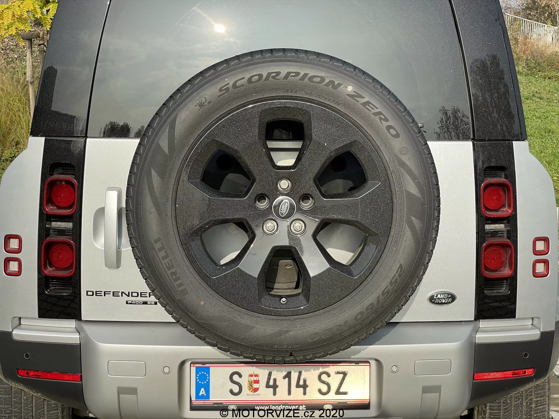 Widok tylny srebrnego Land Rovera Defendera 2020 z widocznym kołem zapasowym, tylnymi światłami, tablicą rejestracyjną, nazwą modelu DEFENDER i znaczkiem Land Rover, srebrnym zderzakiem z czujnikami i kamerą oraz odblaskami. SUV jest zaparkowany w świetle dziennym, co podkreśla czysty i dobrze utrzymany stan pojazdu.