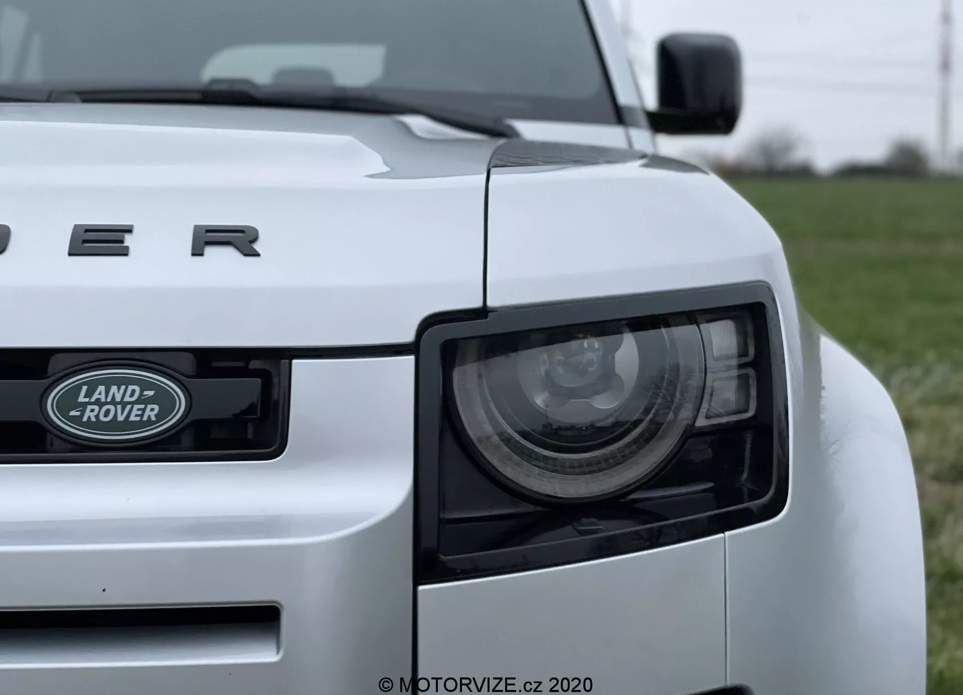 Zbliżenie białego samochodu Land Rover Defender 2020 wykonane z niskiego kąta, przedstawiające lewy przedni kwartał pojazdu. Charakterystyczne cechy to reflektor, kratka z emblematem 'LAND ROVER' oraz maska z napisem 'DEFENDER'. Reflektor zawiera nowoczesne elementy LED, a solidny design samochodu jest podkreślony widocznymi liniami i krzywiznami. Samochód jest umieszczony na trawiastym polu, z rozmytym tłem, co podkreśla pojazd.