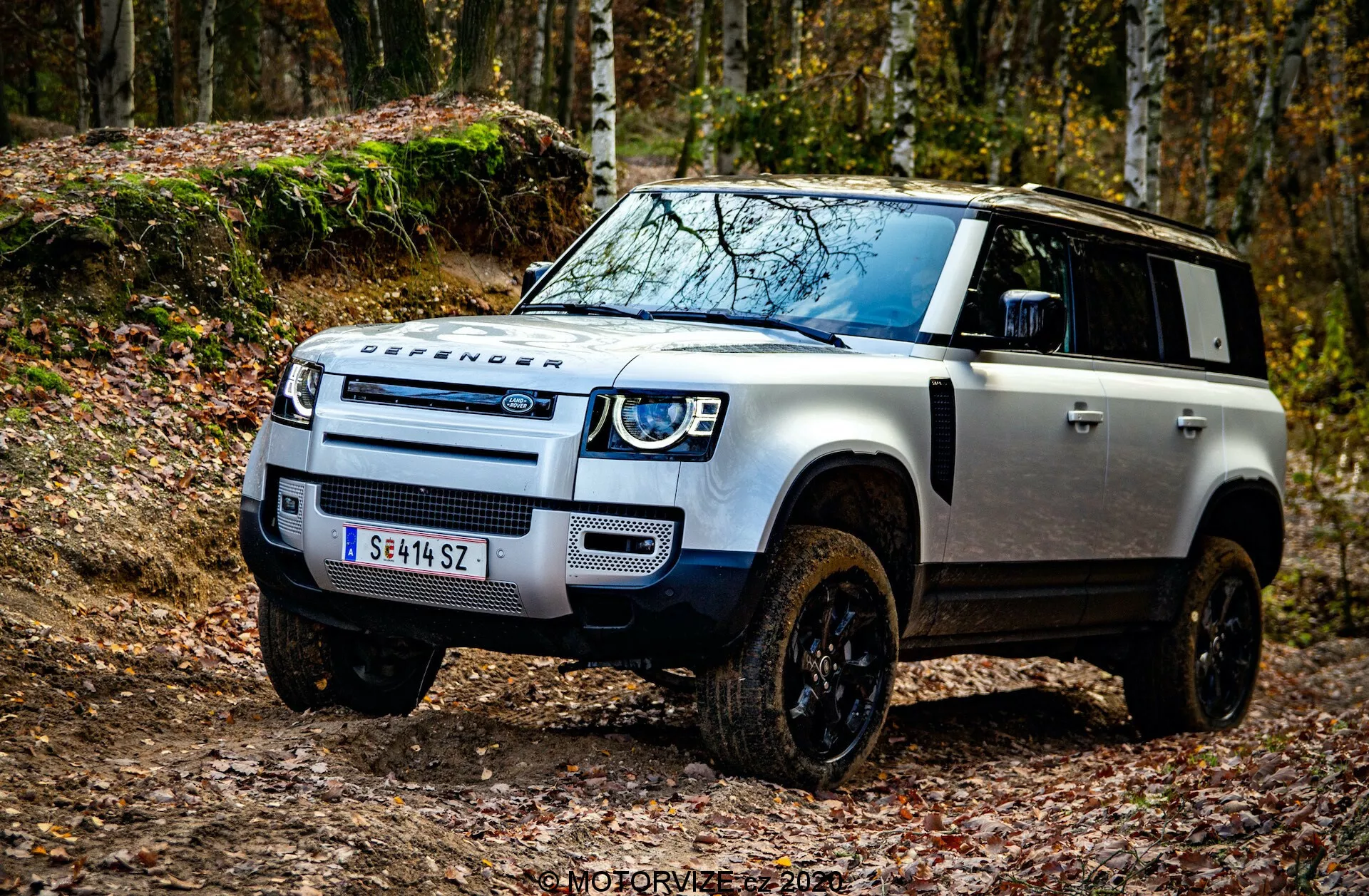 Zdjęcie z niskiego kąta uchwycone zostało Land Rover Defender 2020 z przedniej lewej części, podkreślając takie cechy terenowe jak charakterystyczna przednia krata Land Rover, reflektory LED z dziennymi światłami, płyta poślizgowa zderzaka przedniego, wyraźne oznaczenie DEFENDER na masce, kontrastowa czarna maska na białym nadwoziu, podniesione wloty powietrza do głębokiej wody, duże nadkola z czarnymi kołami terenowymi, kierunkowskazy na lusterkach bocznych i zaawansowana technologia wspomagania kierowcy w pobliżu przedniego zderzaka. Obrazek umieszczony jest w terenowym otoczeniu z opadłymi liśćmi, podkreślając przygodowy wygląd pojazdu. Znacznik wodny 'MOTORVIZA 2021'.