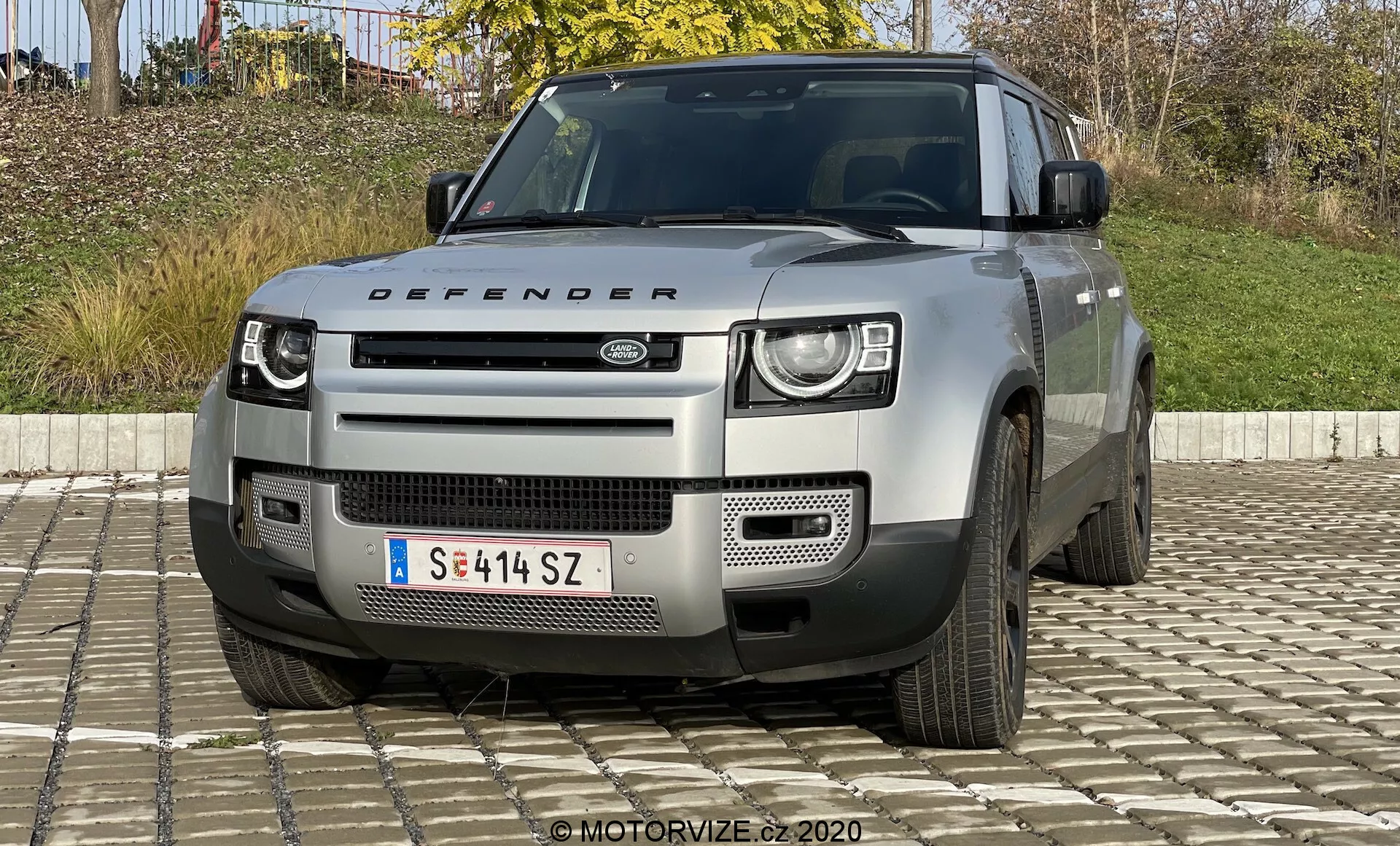 Widok od przodu modelu Land Rover Defender 2020, podkreślający jego charakterystyczne dwupoziomowe reflektory LED, wyraźną kratkę z logo Land Rover, geometryczny zderzak z srebrną płytą ochronną oraz inne solidne cechy sugerujące zdolności terenowe. Widzoczne są również pokrywa silnika z napisem 'Defender', wloty powietrza i częściowy widok pięcioramiennych kół. Pojazd w jasnym neutralnym kolorze, prawdopodobnie srebrnym lub szarym.