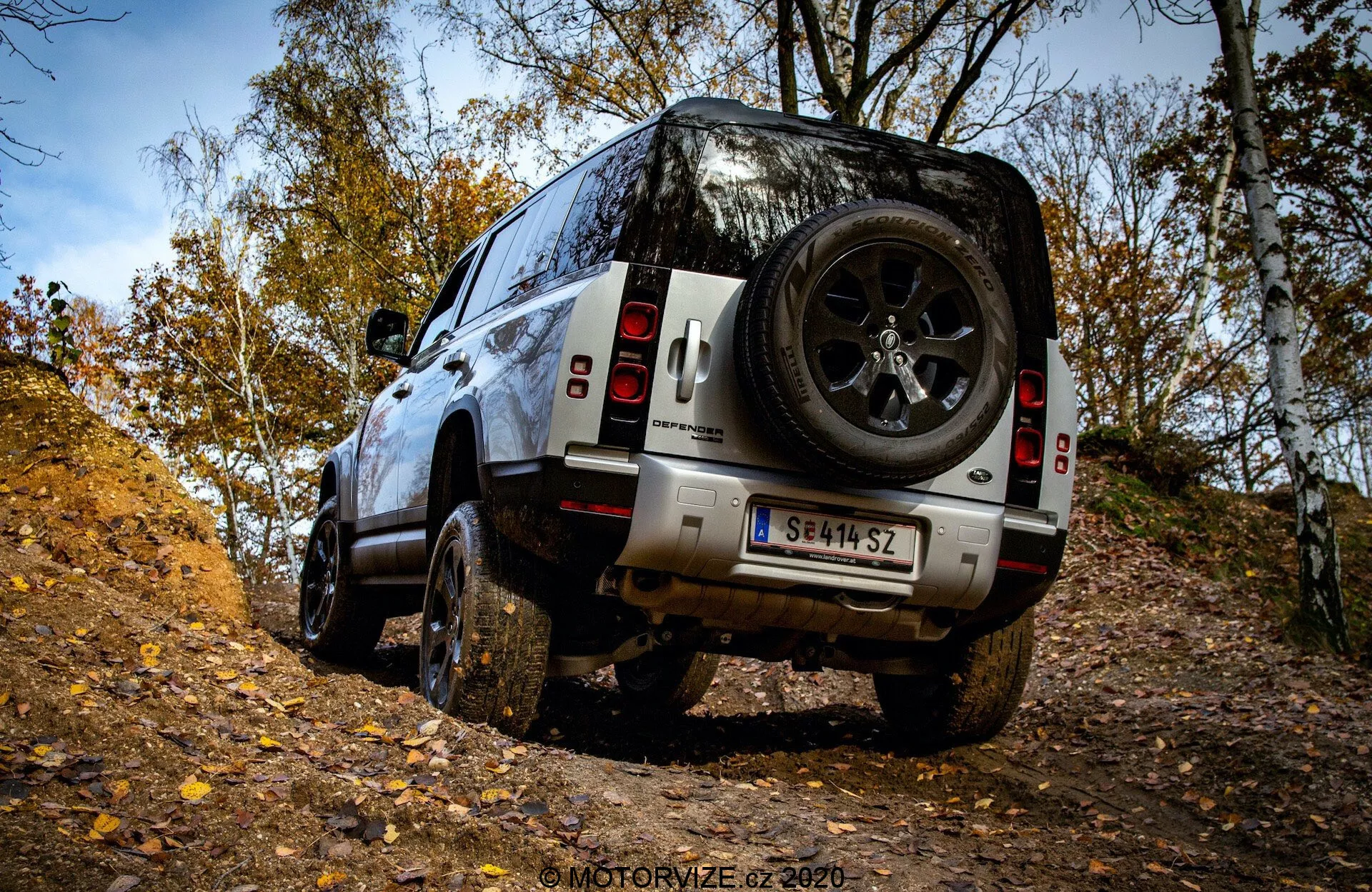 Widok tylno-tylny w trzech czwartych SUV-a Land Rover Defender 2020 na trudnym terenie, prezentującego wytrzymałe opony, dużą wysokość podwozia i tylne koło zapasowe w jesiennym lesie.