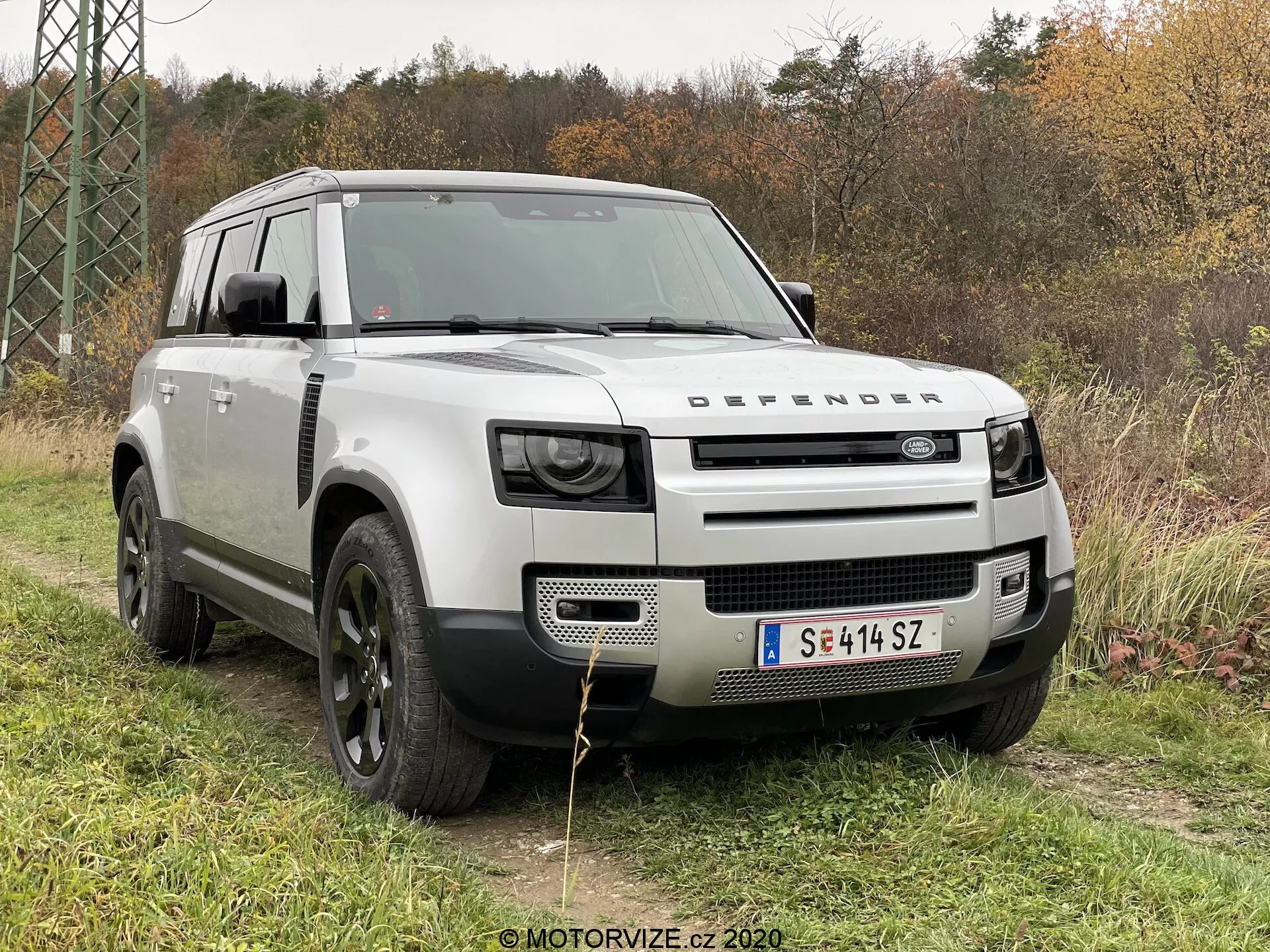 Ten obraz przedstawia Land Rover Defender 2020 w scenerii plenerowej na trawie z listowiem i drzewami w tle. Uchwycony z kąta trzech czwartych z przodu, zdjęcie podkreśla wyraźny grill z napisem 'DEFENDER', nowoczesne reflektory LED, solidną maskę z projektem muszli, duże czarne koła aluminiowe z terenowymi oponami oraz nadwozie w jasnosrebrnym kolorze kontrastującym z czarnymi nadkolami i dachem. Elementy designu, takie jak przednia płyta ochronna, wloty powietrza 'alpine' i kwadratowa konstrukcja podkreślają jego zdolności terenowe i użytkowe.
