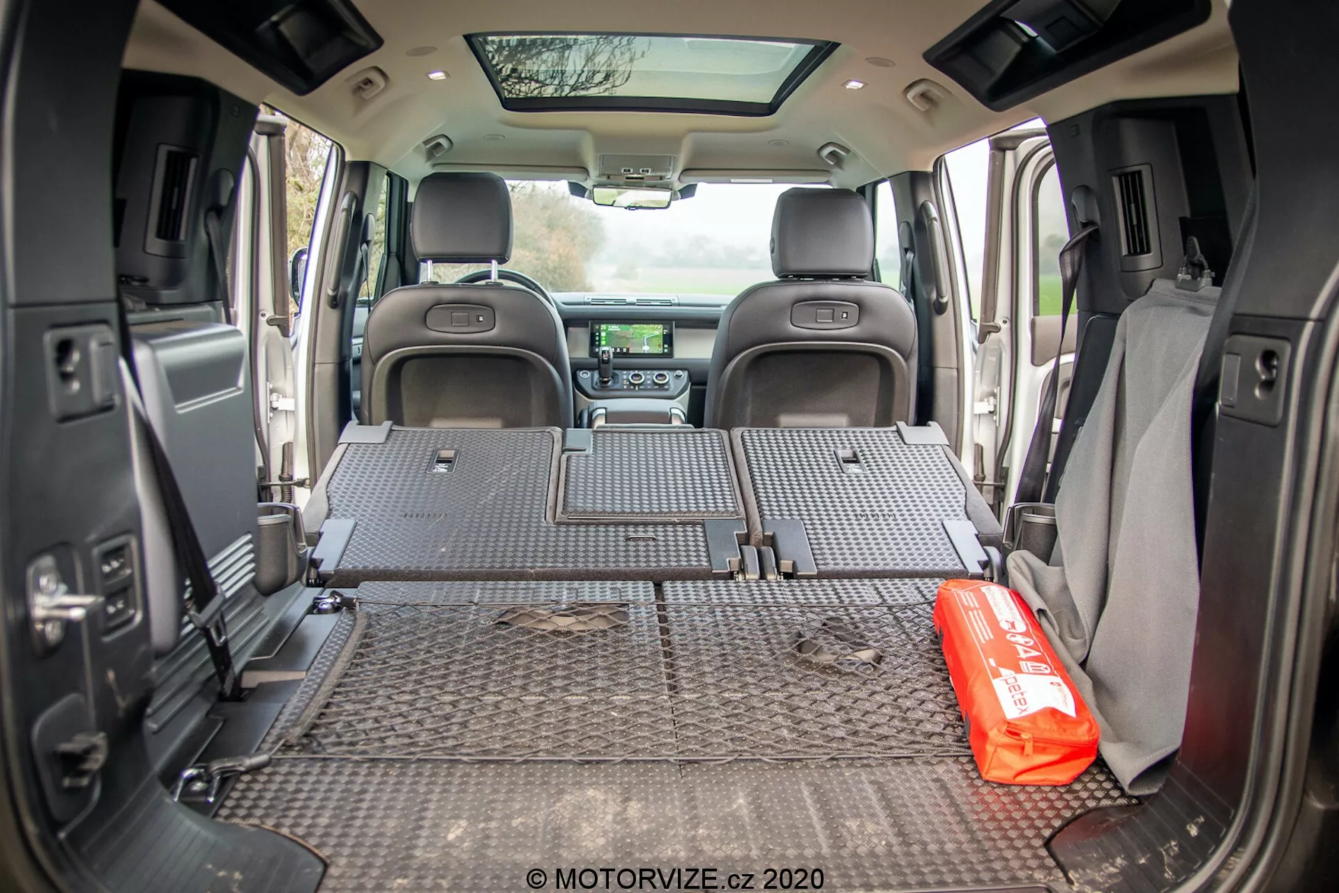 Widok wnętrza przestrzeni ładunkowej Land Rover Defender 2020 wykonany z tylnej perspektywy, pokazujący ochronną matę z wzorem diamentowej blachy, pokrywającą całą podłogę bagażnika. Tylne siedzenia są złożone na płasko, odsłaniając przestronną przestrzeń bagażową. Ściany boczne mają różne elementy, takie jak głośniki i uchwyty na kubki, co sugeruje zrównoważony projekt między funkcjonalnością a komfortem. Odległa deska rozdzielcza z przodu jest częściowo widoczna, zawierając system infotainment i sterowanie klimatyzacją. Na macie ładunkowej jest wyraźnie umieszczona czerwona apteczka, co sugeruje rodzinne lub przygodowe zastosowanie pojazdu. Otwarta tylna klapa pozwala, aby naturalne światło wpadało do przestrzeni ładunkowej.
