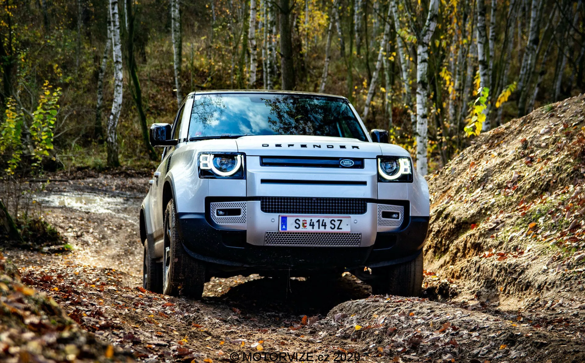 Widok z przodu Land Rovera Defendera 2020, uchwycony z niskiego kąta, podkreślając jego grill, reflektory i przedni zderzak. SUV jest ustawiony na nieutwardzonej drodze z leśnym tłem, co podkreśla jego gotowość do jazdy terenowej. Widoczne elementy obejmują wyraźne oznaczenie 'DEFENDER', reflektory LED, szerokie błotniki i solidne opony.