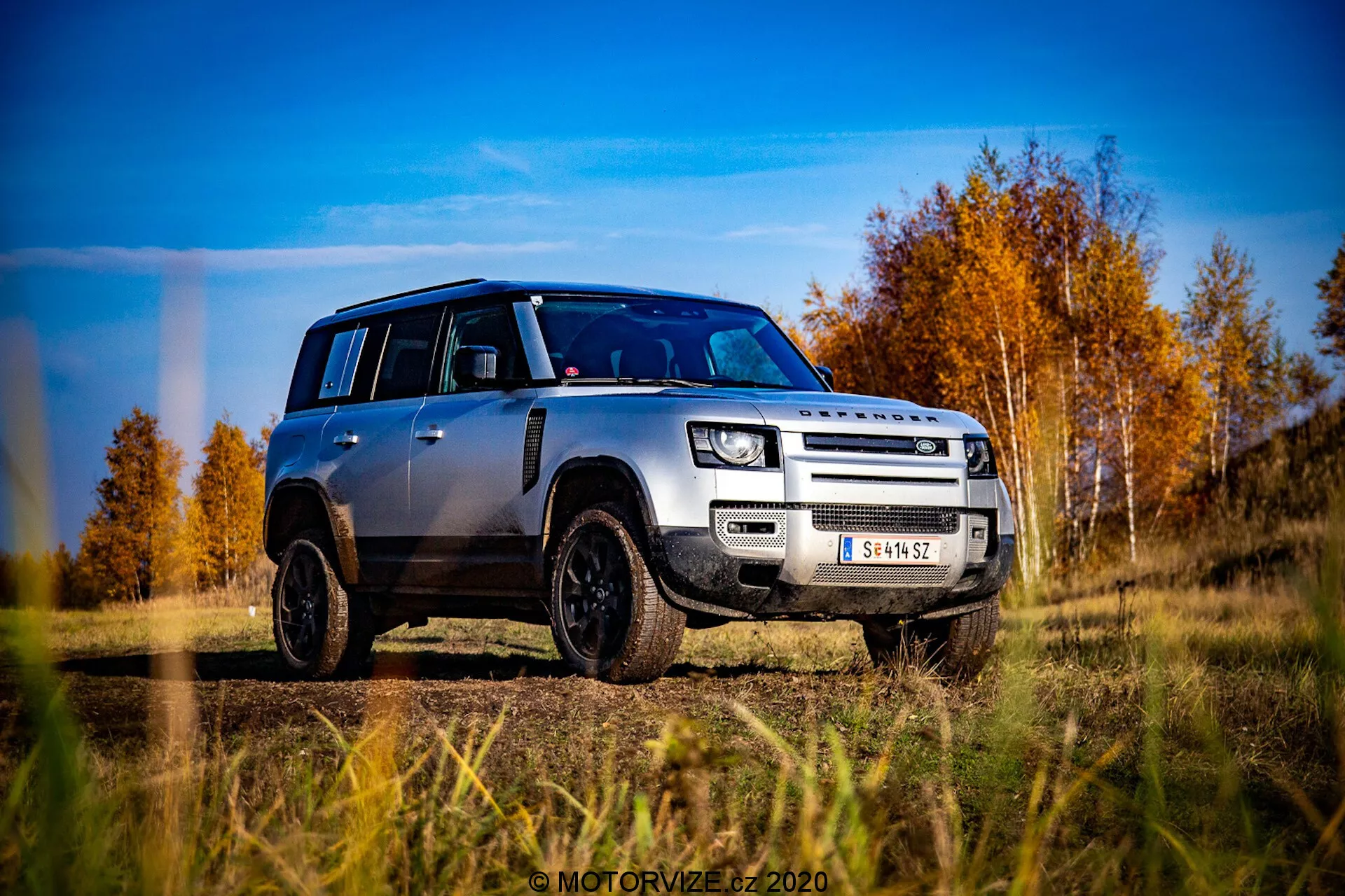 Obraz srebrnego Land Rover Defender 2020 widzianego z perspektywy poziomu gruntu, uchwycony w niskim, ukośnym, trzykwartersowym widoku od przodu. Pojazd wyraźnie pokazuje swoją kratkę, maskę, przedni zderzak i design kół, lekko skierowany w stronę kierowcy. Defender znajduje się na terenie zewnętrznym z tłem jesiennych drzew o czerwonych i pomarańczowych liściach pod częściowo zachmurzonym niebem. Charakterystyczne cechy projektowe obejmują charakterystyczny pudełkowy kształt, nowoczesne reflektory LED, światła przeciwmgielne, kratkę z dwoma poprzeczkami z napisem LAND ROVER, przednią osłonę, czarne felgi ze stopów z oponami terenowymi i kontrastujący czarny dach z pasującymi czarnymi obudowami lusterek. Zdjęcie jest bardzo ostre i dynamiczne, podkreślając wytrzymałość i nowoczesną estetykę pojazdu.