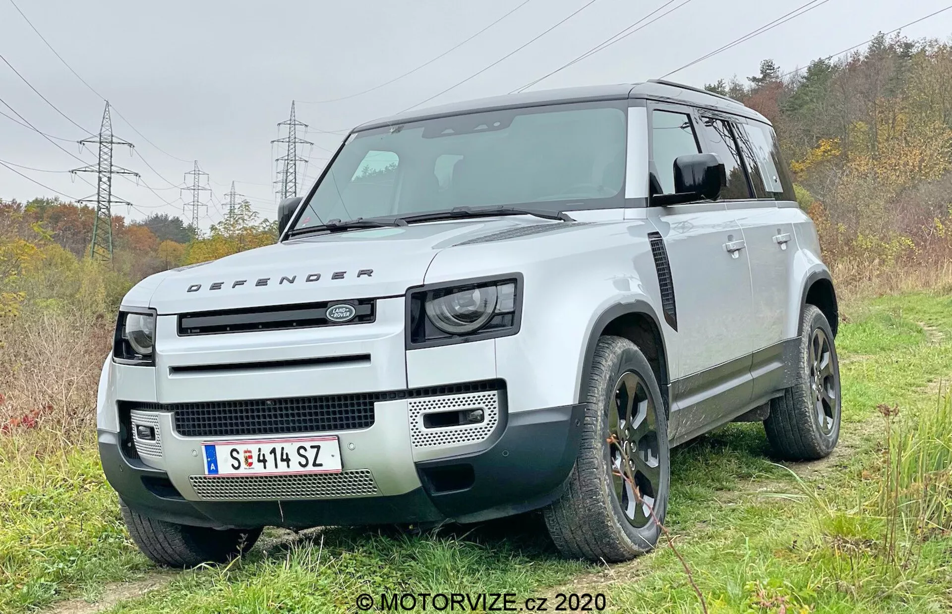 Ta fotografia przedstawia nowoczesny SUV Land Rover Defender 2020 z kąta przedniego ćwierćboku, pokazując stronę kierowcy. Pojazd jest lekko poza drogą na trawiastym obszarze z widoczną ścieżką z ziemi, podkreślając jego zdolności terenowe. Charakterystyczny kwadratowy design posiada dużą prześwitowość, dwukolorowy schemat z białym nadwoziem i kontrastującym czarnym dachem. Przednia część ukazuje reflektory LED zainspirowane retro, maskę z emblematem Defendera oraz minimalistyczny przedni grill, podkreślając jego reputację wytrzymałego pojazdu terenowego. Widoczne cechy techniczne obejmują przednie czujniki parkowania zintegrowane z zderzakiem, sugerując zaawansowane systemy wspomagania kierowcy. Solidne nadkole pomieszczą duże terenowe opony, a takie elementy jak felgi aluminowe i unikalny design grilla są wyróżnione.