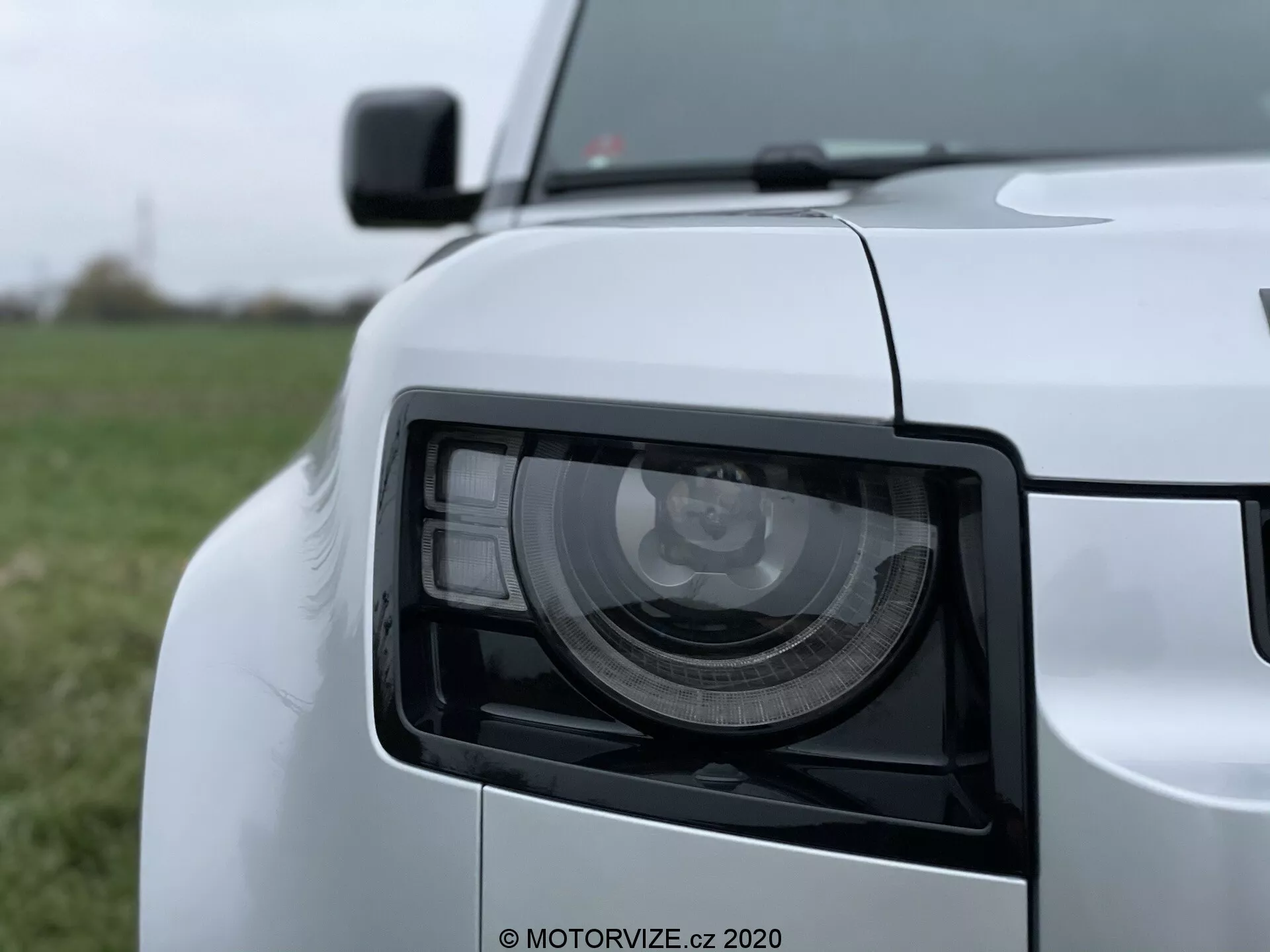 Zbliżenie na przedni lewy reflektor Land Rover Defender 2020 z nowoczesnym, kanciastym designem, wykonane pod lekkim kątem z perspektywy niskiej do średniej wysokości. Na obrazie widoczne są szczegółowe moduły świetlne LED/HID, błyszczące białe wykończenie lakieru oraz nieostre fragmenty przedniego grilla, krawędzi maski, przedniej części zderzaka oraz lusterka bocznego.