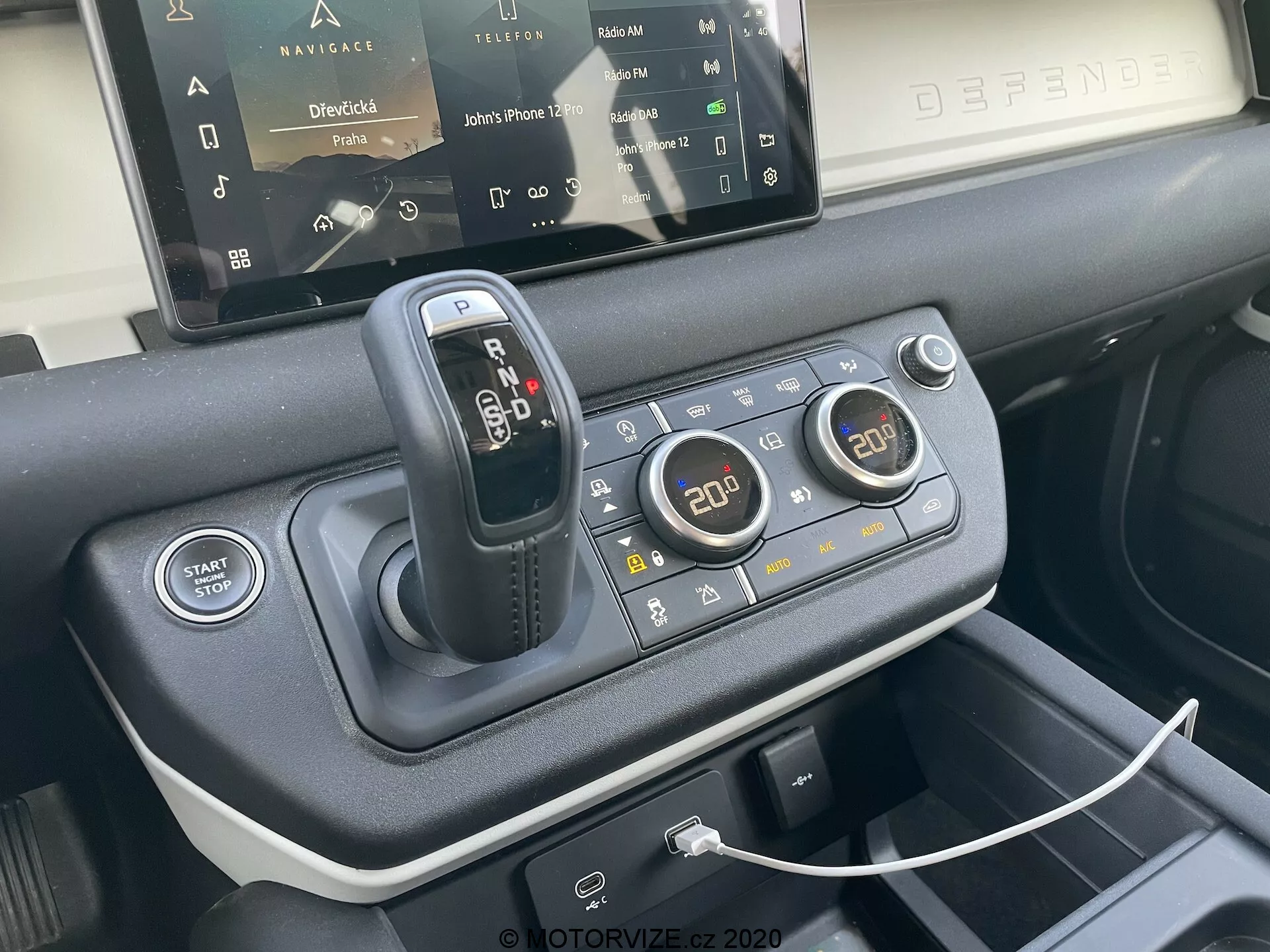 Zbliżenie konsoli centralnej Land Rover Defender 2020, podkreślającej dźwignię zmiany biegów, system reagowania na teren dla różnych warunków jazdy, przycisk start/stop silnika, dwustrefowe sterowanie klimatyzacją, port USB do podłączenia urządzeń, częściowo widoczny system infotainment z ekranem dotykowym oraz materiały premium użyte w projekcie wnętrza, widziane z perspektywy przedniego siedzenia pod kątem 45 stopni.