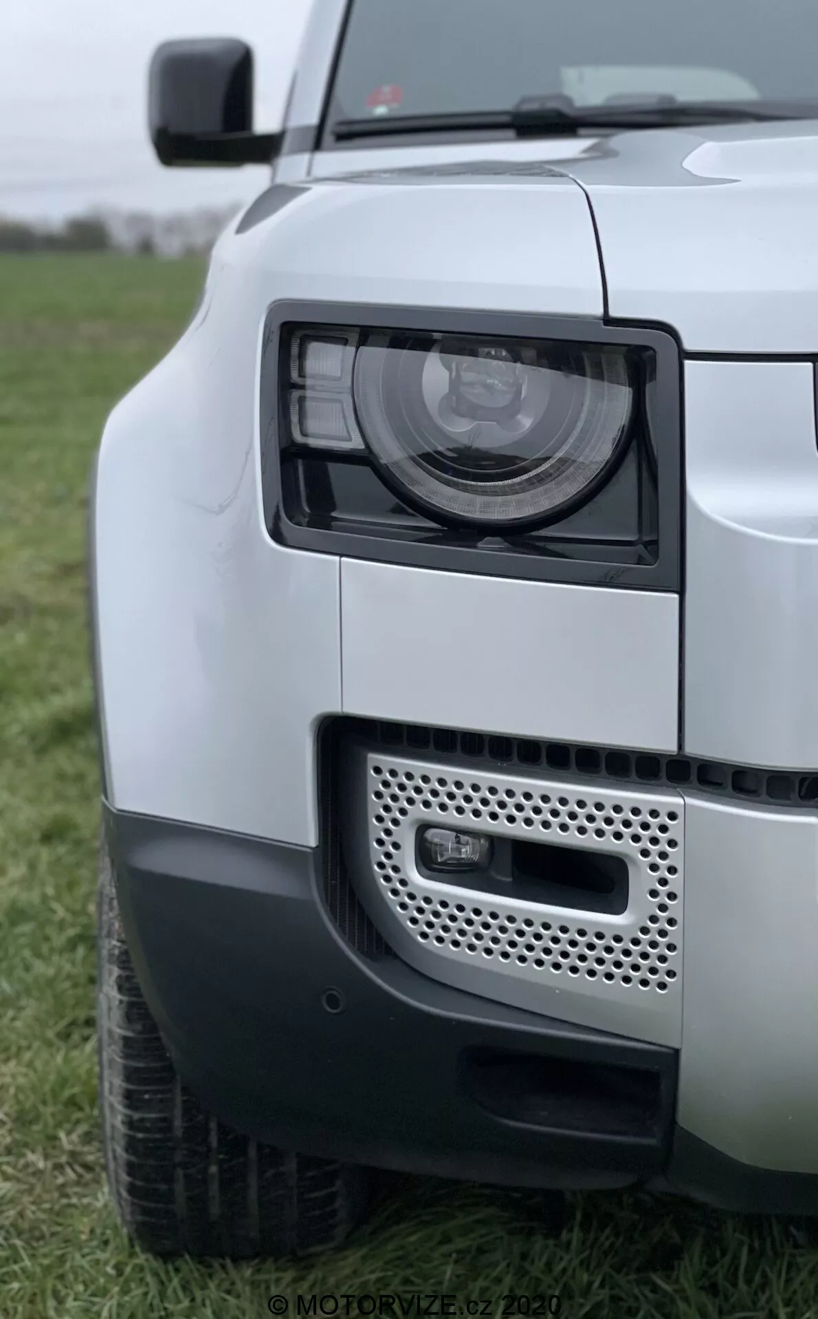Zbliżenie na Land Rover Defender 2020 z naciskiem na zespół reflektorów, kratkę i przedni błotnik. Zdj��cie pokazuje pojazd z przedniego kąta bocznego, ukazując nowoczesne reflektory LED/HID, adaptacyjną technologię oświetlenia, kratkę z możliwym umiejscowieniem czujników oraz dwutonowy schemat malowania. Przedni zderzak prezentuje światło przeciwmgielne i system czujników parkowania, podkreślając solidny i nowoczesny design SUV-a Defendera.