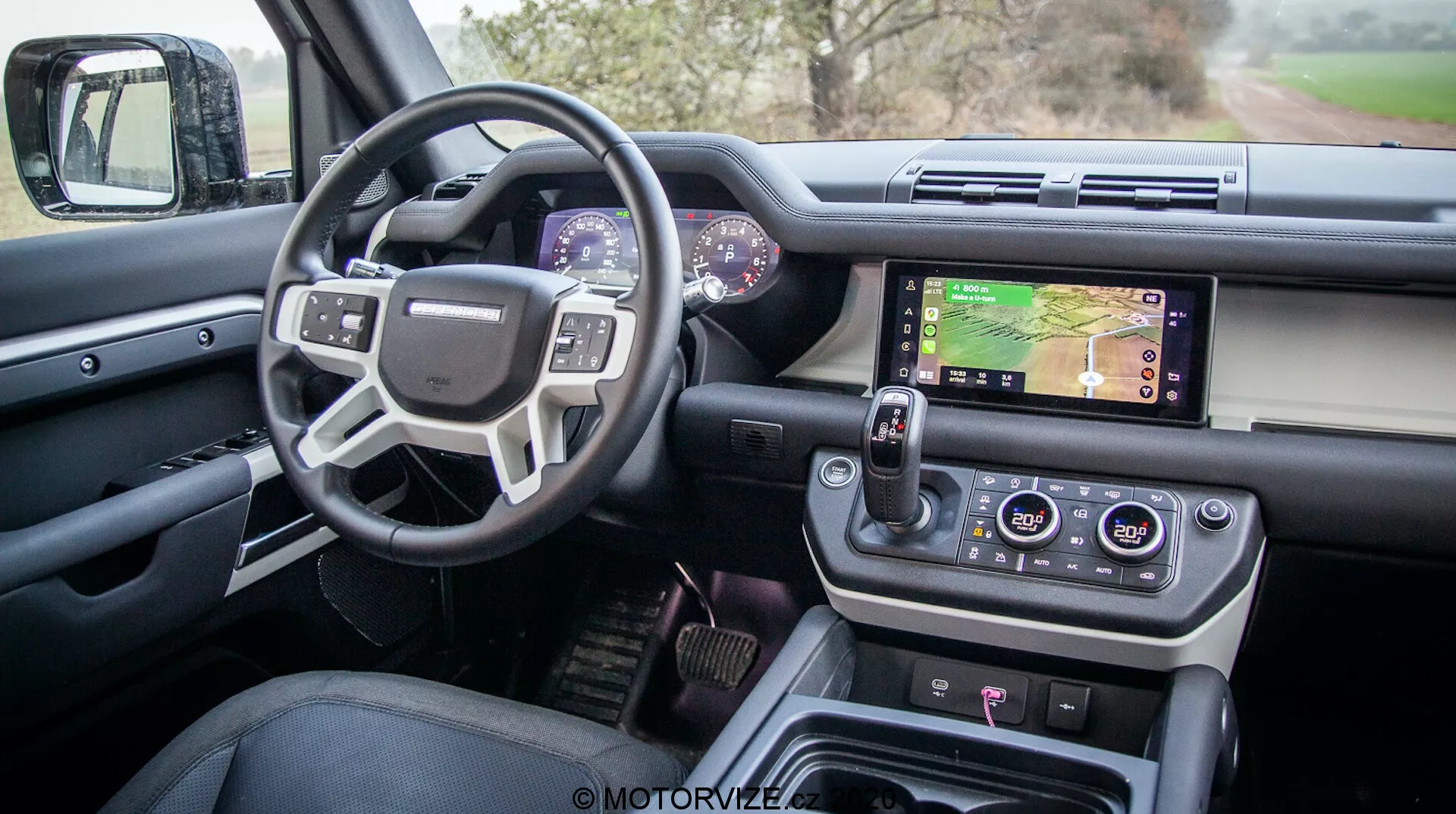 Wnętrze nowoczesnego Land Rovera Defender 2020 widziane z drzwi kierowcy, ukazujące zaawansowany design i funkcje, w tym otwarte drzwi kierowcy z przyciskami do regulacji szyb i lusterek, wielofunkcyjną kierownicę z przyciskami kontrolnymi, cyfrowy zestaw wskaźników, centralny system infotainment z ekranem dotykowym wyświetlającym mapę, pokrętła klimatyzacji, nowoczesny obrotowy selektor biegów i przyciski kontrolne do wyboru trybu jazdy, wszystko w luksusowym wykończeniu wysokiej jakości.