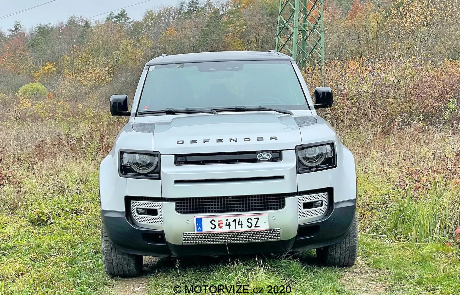 Widok z przodu białego Land Rover Defender 2020 4x4 z wyraźnym prostokątnym grillem otoczonym okrągłymi reflektorami LED z zintegrowanymi kwadratowymi światłami do jazdy dziennej, solidnym przednim zderzakiem z prostokątnymi otworami, płaską maską z napisem 'LAND ROVER' i oznaczeniem 'DEFENDER' na przedniej krawędzi, płaską szybą przednią, centralnie zamontowaną europejską tablicą rejestracyjną i czujnikami parkowania, sfotografowane nieruchome w naturalnym otoczeniu.