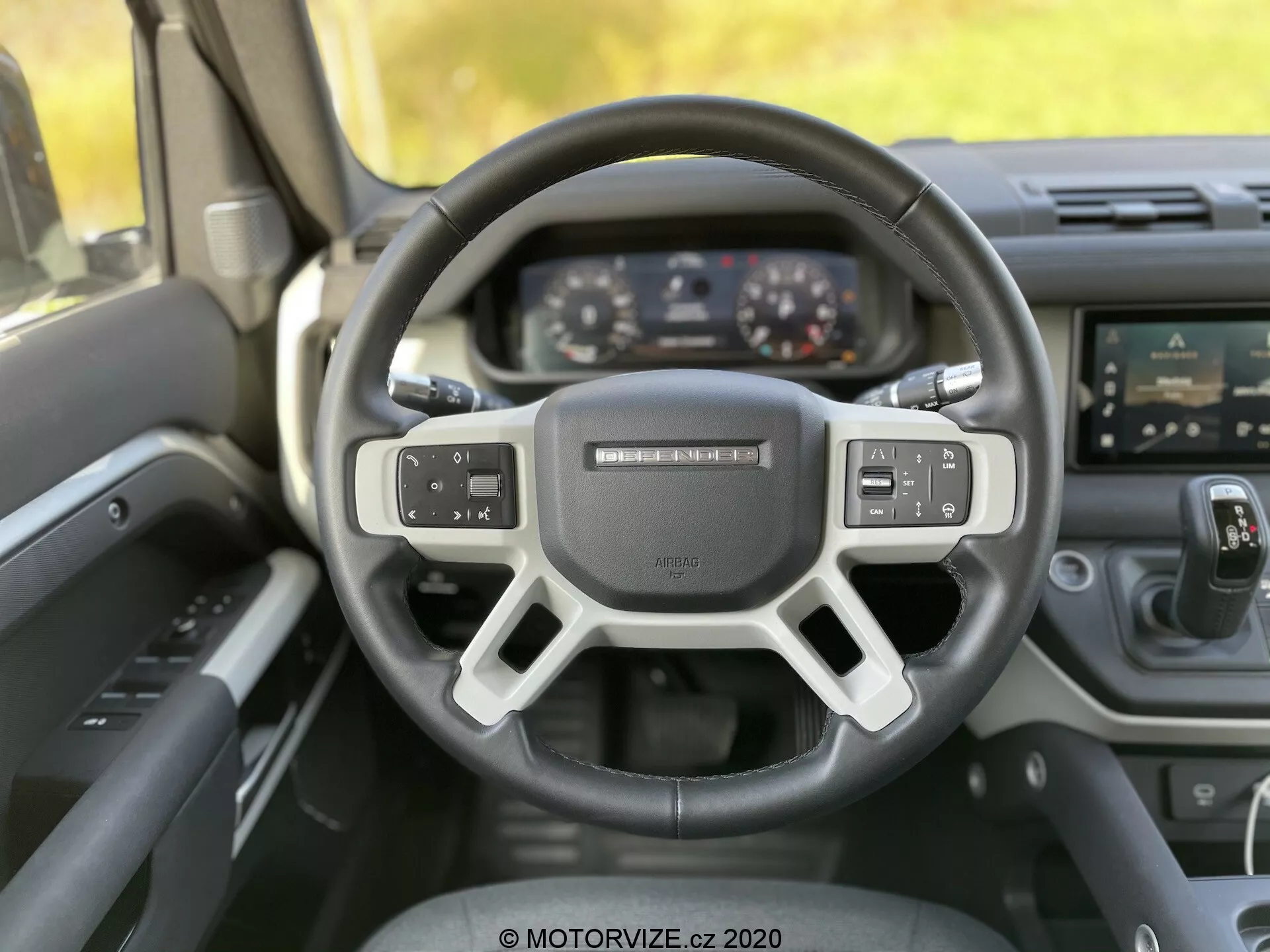 Widok wnętrza Land-Rover Defender 2020 z perspektywy kierowcy, ukazujący nowoczesną, trójramienną kierownicę z zintegrowanymi kontrolkami do audio, poleceń głosowych i ustawień tempomatu. Za kierownicą znajduje się cyfrowy panel instrumentów z wyświetlaczami prędkościomierza, obrotomierza oraz wielofunkcyjnego ekranu. Obraz przedstawia również konsolę centralną z dotykowym systemem infotainment i przyciskami klimatyzacji poniżej. Kąt fotografii jest nieco przechylony w górę, podkreślając ergonomiczny design, zaawansowaną technologię i materiały premium nieruchomego samochodu.