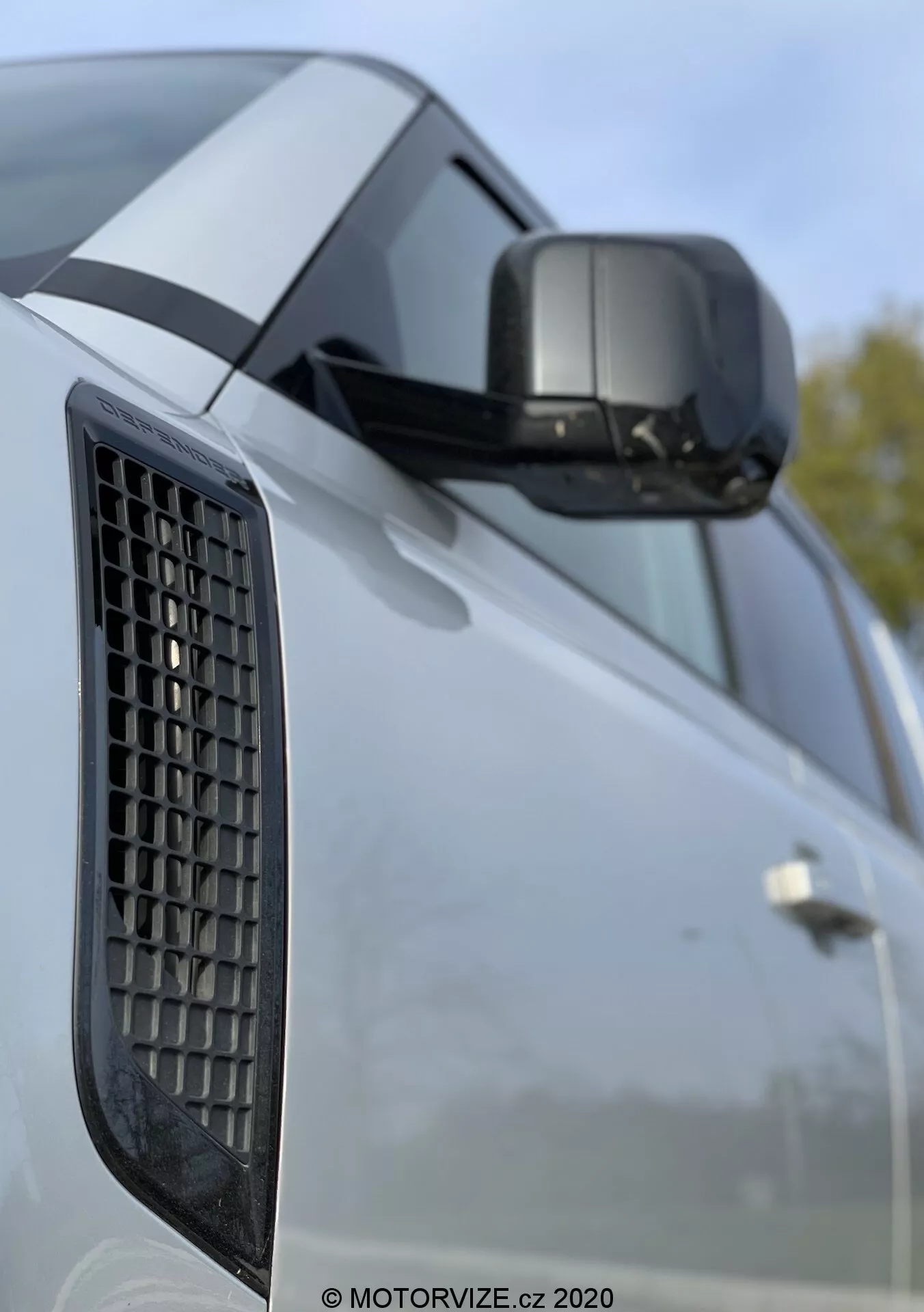 Zbliżenie na Land Rover Defender 2020 z uwypukleniem błotnika bocznego z wentylatorem, bocznego lusterka z wbudowanym kierunkowskazem i fragmentu drzwi z wyraźnymi liniami charakterystycznymi; zdjęcie skupia się na elementach designu pojazdu z rozmytym tłem sugerującym zewnętrzne otoczenie.