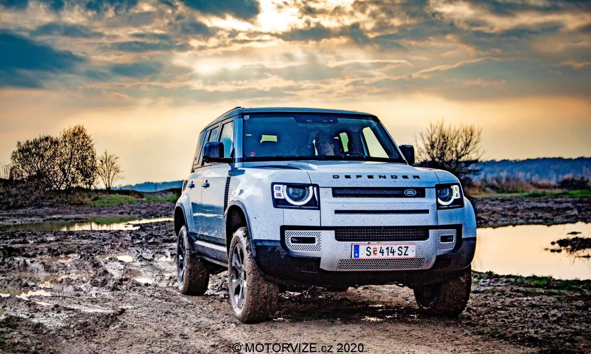 Fotografia SUV Land Rover Defender wykonana z przedniego, lekko nieosiowego kąta, ukazująca przednią część pojazdu i jego zdolność terenową. Pojazd jest w jasnym kolorze, z brudem na oponach i dolnych panelach nadwozia, co wskazuje na jazdę terenową. Samochód wyposażony jest w snorkel do brodzenia w głębokiej wodzie i reflektory LED. Wiejskie otoczenie obejmuje błotnisty teren i nieruchome lustro wody, z dramatycznym niebem wczesnego wieczoru w tle.