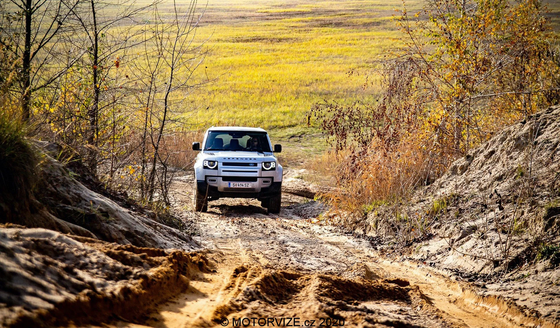SUV Land Rover Defender 2020 jest widoczny z przodu podczas jazdy terenowej, umieszczony centralnie na tle nierównego terenu i naturalnego otoczenia. Widoczna przednia część samochodu prezentuje zaawansowaną artykulację zawieszenia, reflektory LED i charakterystyczny grill z poziomymi listewkami. Uchwycenie SUV-a w wiejskim otoczeniu z jesiennymi odcieniami i naturalnym światłem, perspektywa wzmacnia solidną i przygodową obecność pojazdu.