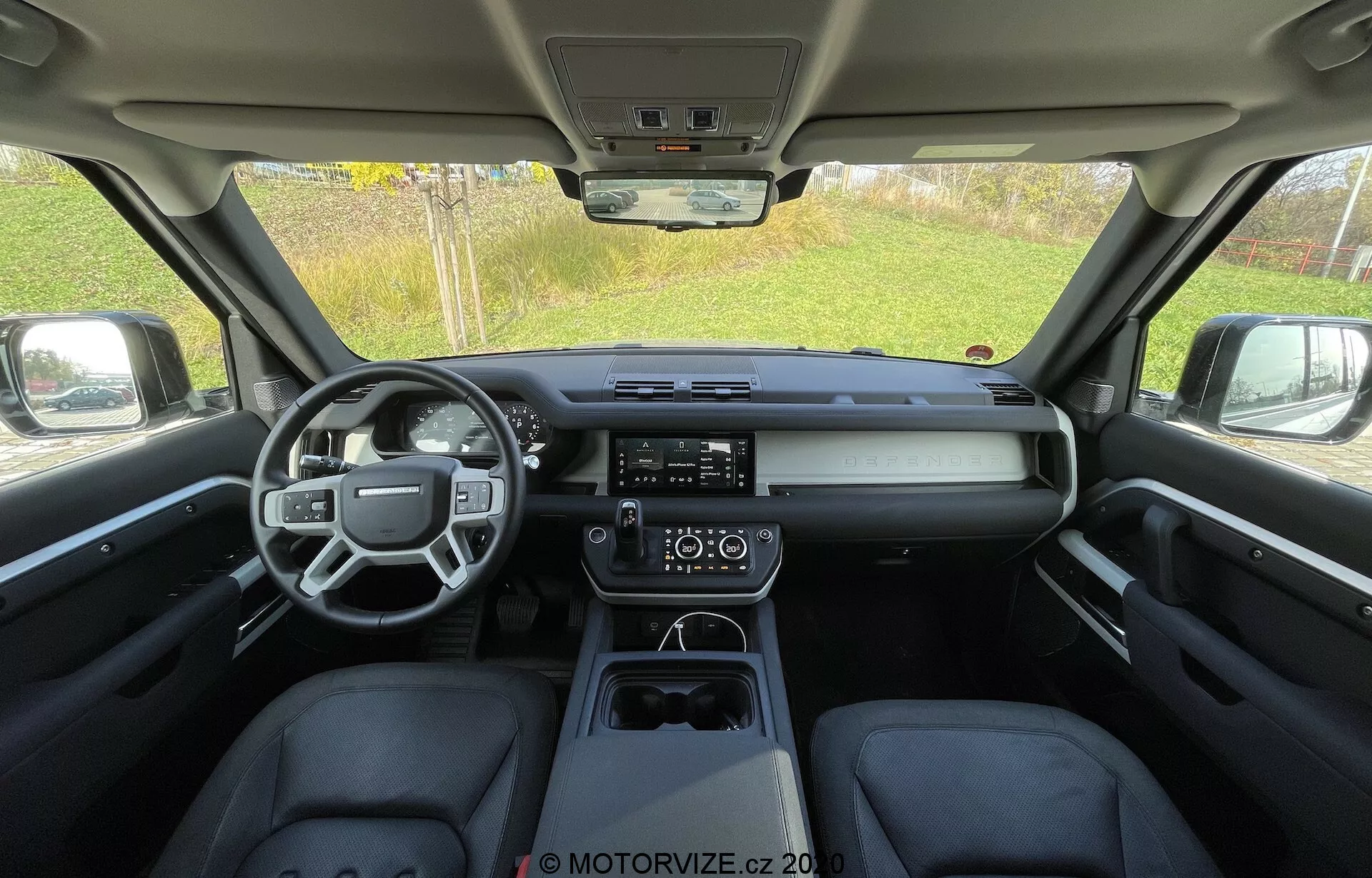Widok wnętrza Land Rover Defender 2020 z tylnego siedzenia, ukazujący elegancką, nowoczesną kabinę z wielofunkcyjną kierownicą, poziomą deską rozdzielczą z centralnym wyświetlaczem cyfrowym, wyraźnym dotykowym systemem multimedialnym, dwustrefową klimatyzacją oraz czystym centralnym panelem z elektronicznym selektorem biegów i uchwytami na kubki.
