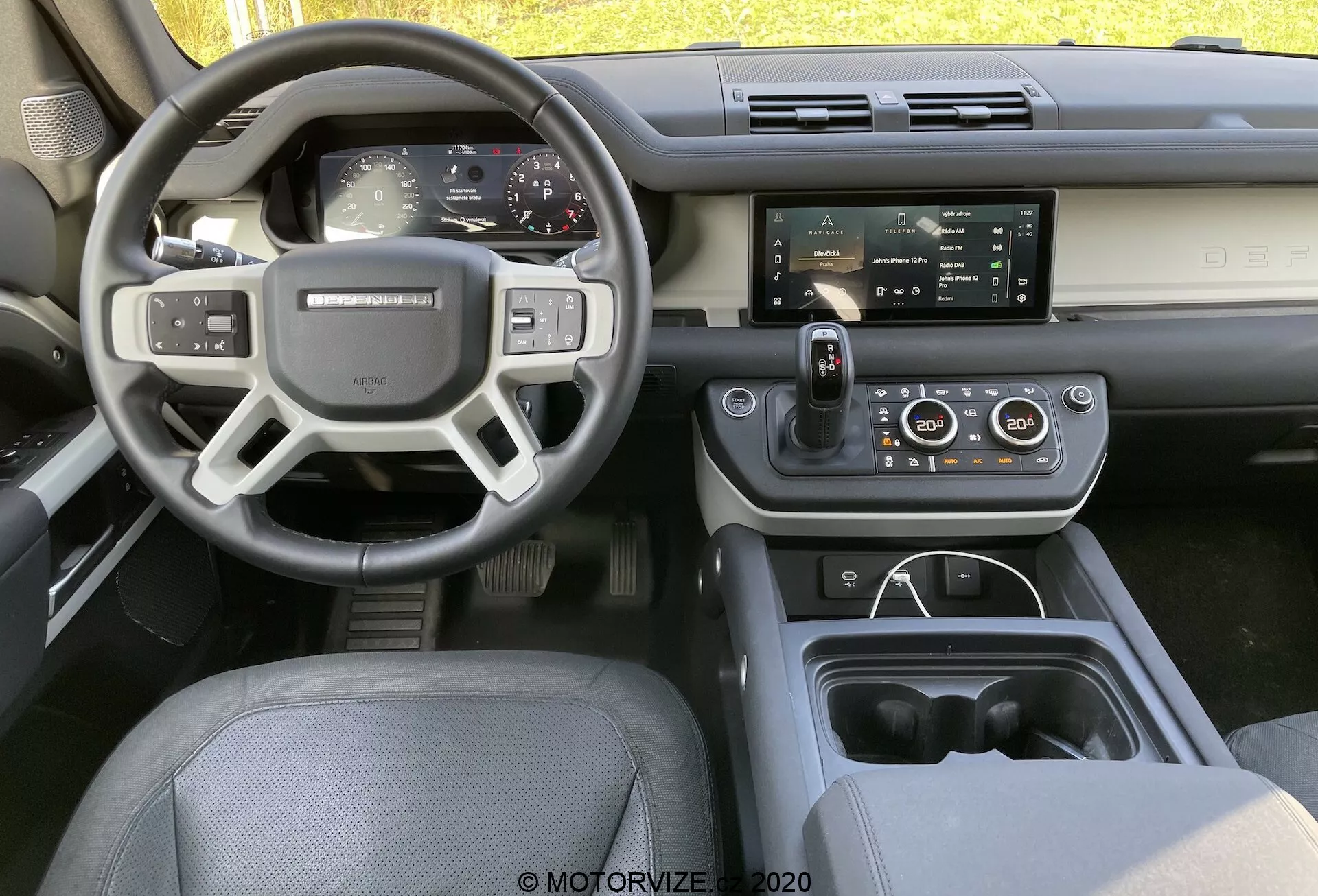 Wnętrze Land Rovera Defender 2020 z tylnego środkowego siedzenia, ukazując deskę rozdzielczą, kierownicę z przyciskami sterującymi, w pełni cyfrowy panel instrumentów i centralny wyświetlacz infotainment z technologiami smart i komfortowymi.