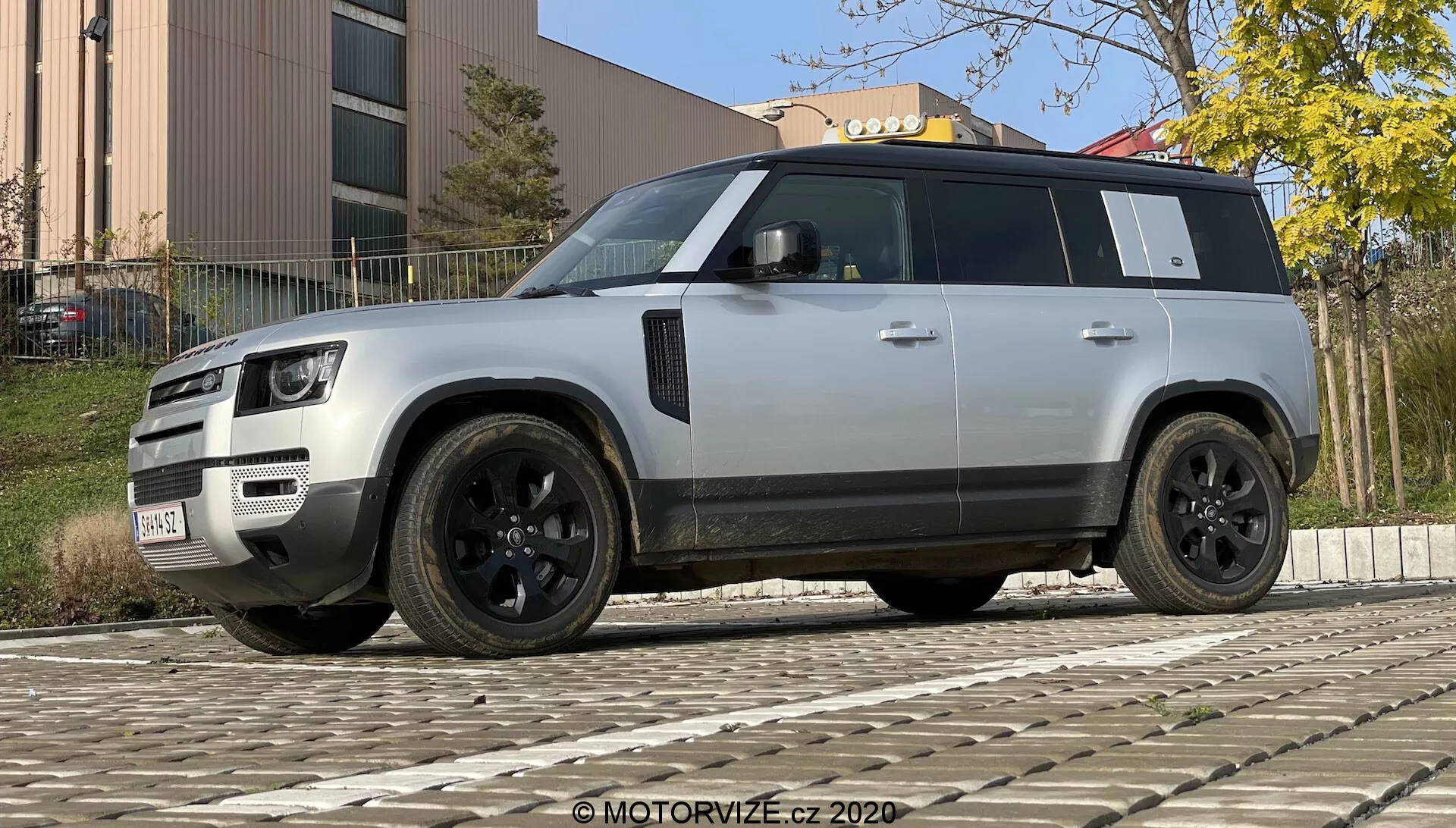 Fotografia nowoczesnego SUV-a Land Rover Defender 2020 wykonana z niskiego kąta, aby podkreślić jego solidną i imponującą postawę. Pojazd jest pokazany w widoku trzy czwarte od przodu i boku, podkreślając jego charakterystyczny grill, reflektory LED, zderzak z lampami przeciwmgielnymi i czujnikami, czarne felgi aluminiowe z wieloszprychowym wzorem i oponami o niskim profilu, czarne lusterka boczne z potencjalnymi dodatkowymi technologiami, przyciemniane szyby, wyraźną linię dachu, chowane klamki drzwi, mocny design nadkoli i jasnosrebrny lub szary kolor kontrastujący z czarnymi elementami wykończenia. SUV jest zaparkowany na bruku, z budynkiem i roślinnością w tle, przy dziennym oświetleniu.