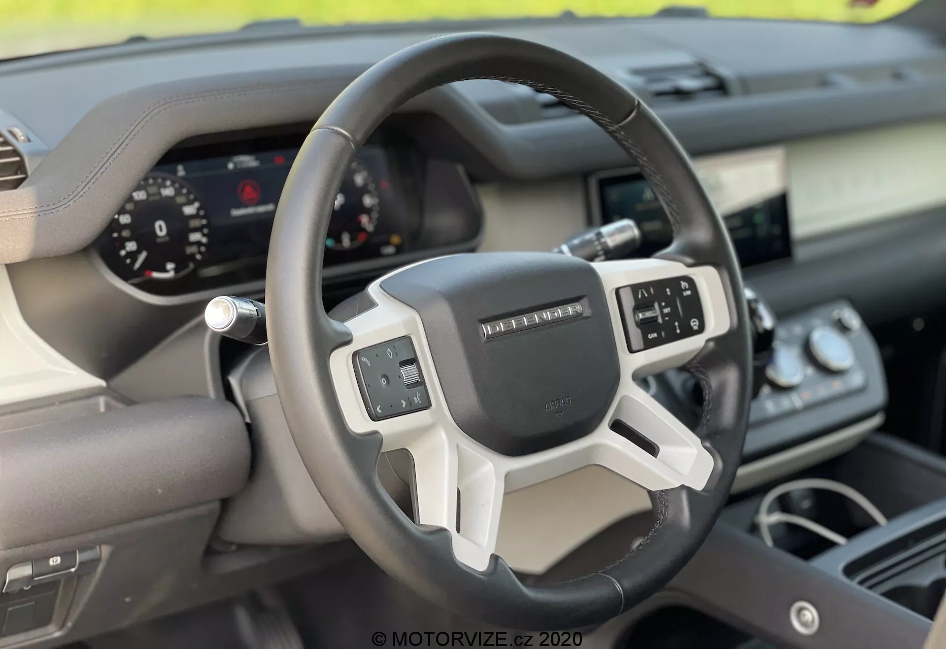 Wnętrze Land Rover Defender 2020, skoncentrowane na kierownicy i desce rozdzielczej z perspektywy przedniego siedzenia pasażera. Nowoczesna kierownica z trzema ramionami posiada wielofunkcyjne sterowanie oraz cyfrowe zestawienie wskaźników dla łatwego odczytu. Mieszanka materiałów, takich jak skóra oraz metalowe lub plastikowe wstawki, podkreśla ergonomiczny i technologiczny design pojazdu.