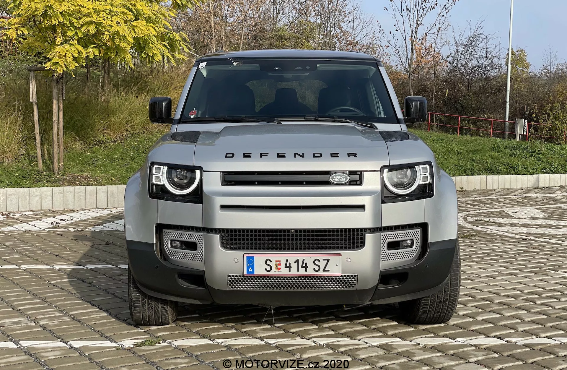 Widok z przodu SUV-a Land Rover Defender 2020, prezentujący charakterystyczną kratkę, reflektory i maskę z napisem 'DEFENDER'. Obraz, centralnie ustawiony i zrobiony pod nieco podwyższonym kątem, podkreśla nowoczesne reflektory LED, geometryczny wzór kratki, przednie czujniki kamer, czujniki parkowania, przednią osłonę i punkty holownicze. Pojazd jest ustawiony na prostym tle z krzewami i poręczą, w naturalnym świetle dziennym.