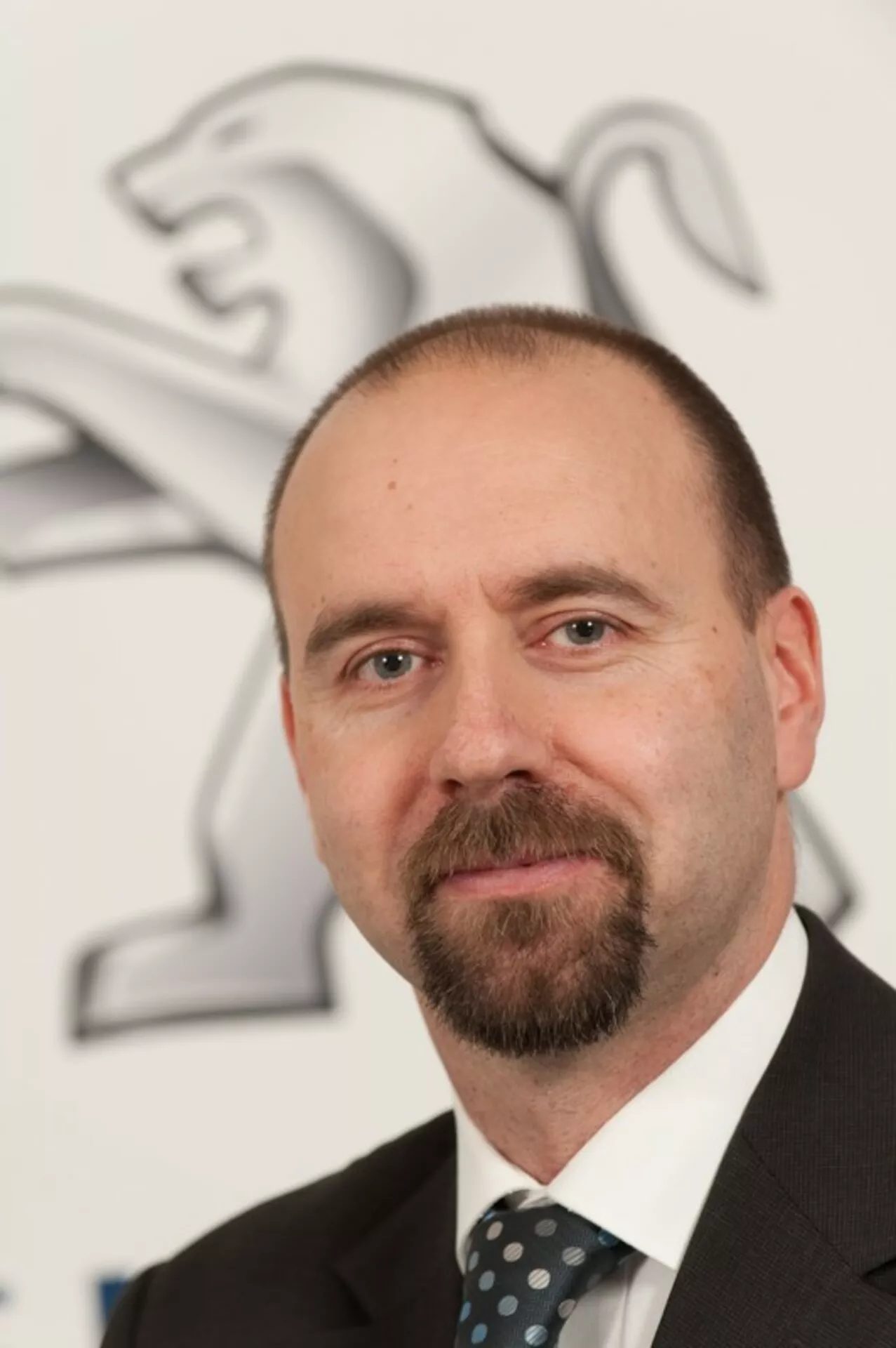 Profesjonalne zdjęcie głowy mężczyzny w garniturze i krawacie z logo Peugeota (lew) w tle, nie pokazuje samochodu