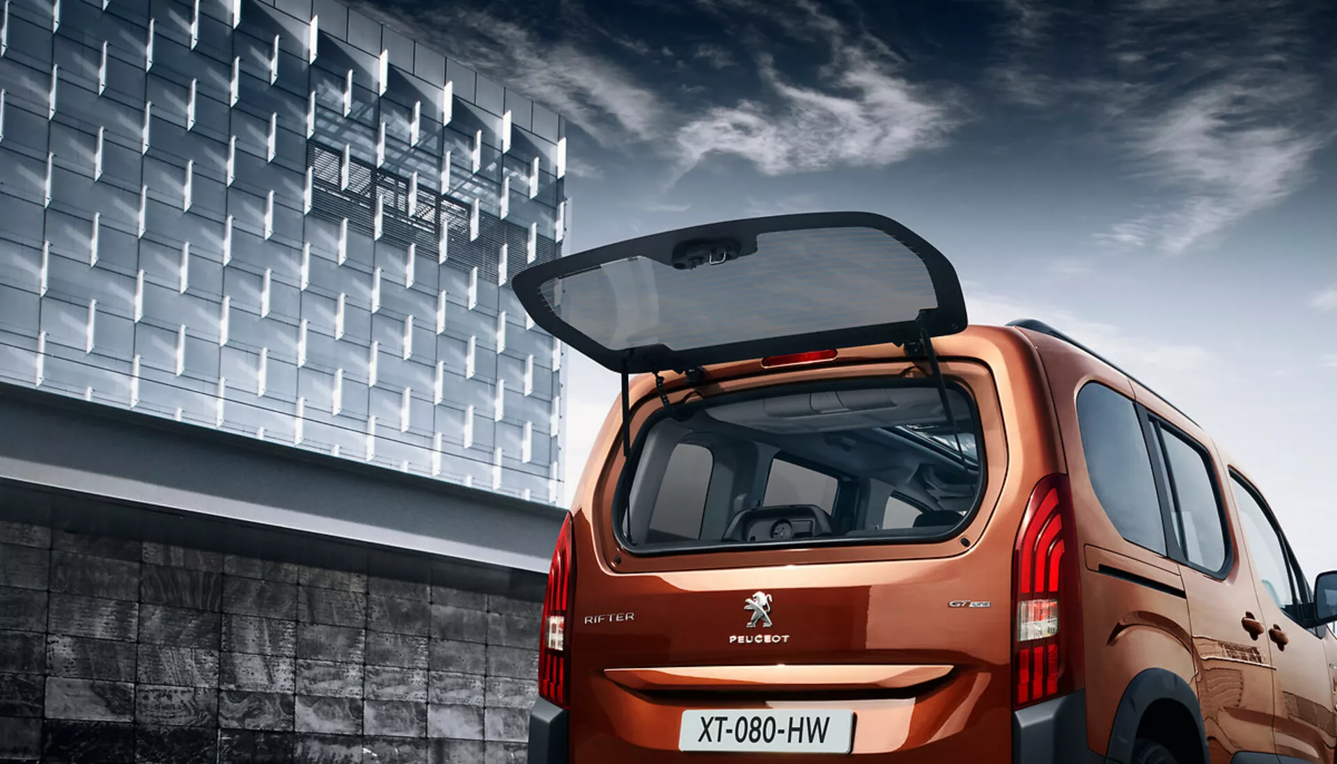 Tylny widok kątowy Peugeota Rifter 2018 ukazujący jego hatchbackowy design, metaliczne wykończenie, otwartą klapę bagażnika z widoczną przestrzenią ładunkową, tylnymi światłami LED i miejskim tłem.