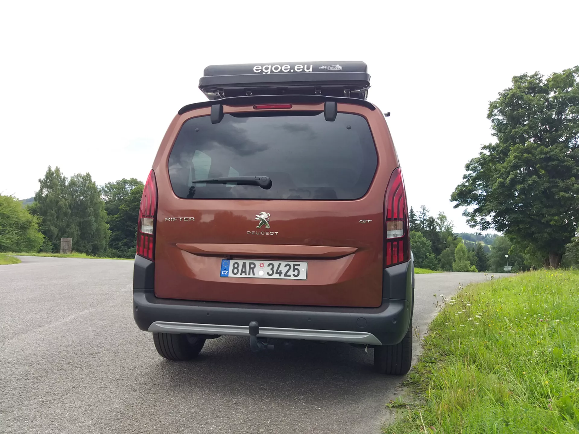 Tylny widok Peugeota Riftera 2018, pokazujący tylną szybę, światła tylne, zderzak i tablicę rejestracyjną, z widocznym logo Peugeota i nazwą modelu Partner. Samochód, zaparkowany na tle natury, wyposażony w box dachowy i posiada akcesorium marki 'egoe.eu', w kolorze czerwonym/bordowym.