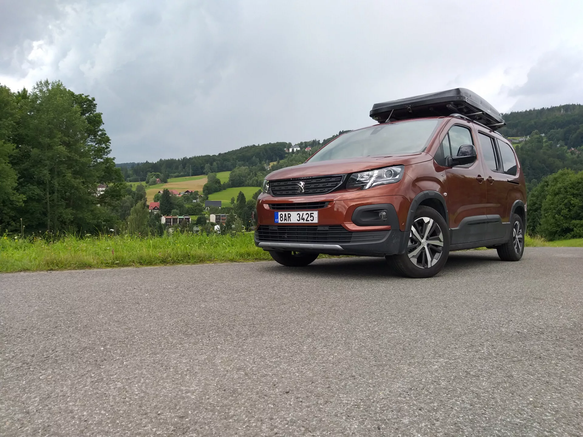 SUV Peugeot Rifter 2018 z metalicznym lakierem i skrzynią dachową, uchwycony z niskiego kąta podkreślający grill, zderzak i felgi aluminiowe, eksponujący zdolności terenowe i nowoczesny design w tym LEDowe reflektory i systemy wspomagania kierowcy ADAS, zaparkowany na utwardzonej nawierzchni na tle krajobrazu wiejskiego