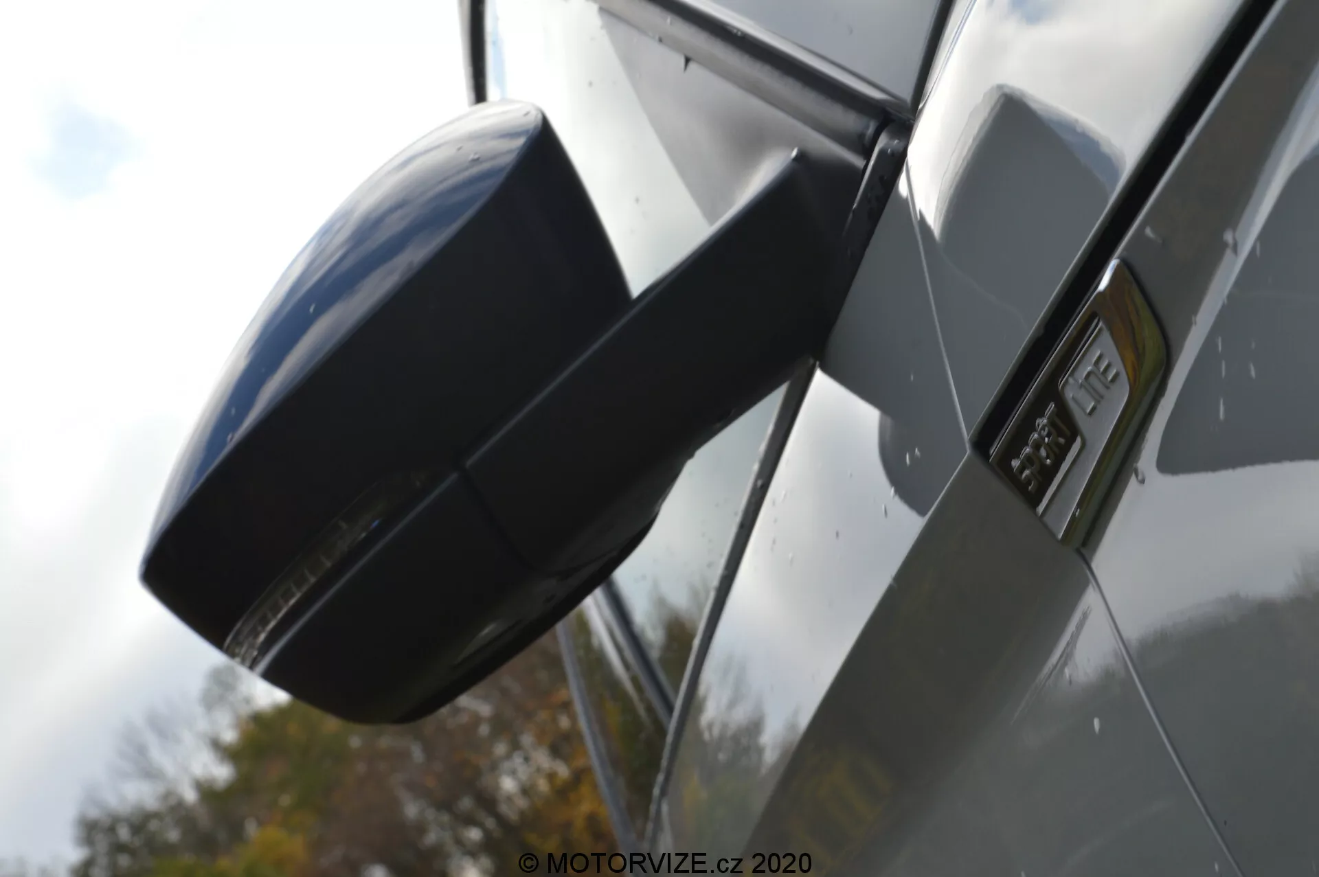 Zbliżony widok boczny Skoda Kodiaq 2017, koncentrujący się na lusterku bocznym i obszarze błotnika. Zdjęcie wykonane pod niskim kątem, podkreślając boczny profil samochodu, z nowoczesną, aerodynamiczną obudową lusterka bocznego w kolorze dopasowanym do nadwozia, wyraźnym oznaczeniem na błotniku wskazującym na konkretny model lub poziom wyposażenia, błyszczącymi metalicznymi panelami nadwozia i odbiciami sugerującymi otoczenie na świeżym powietrzu z ciemnym samochodem.