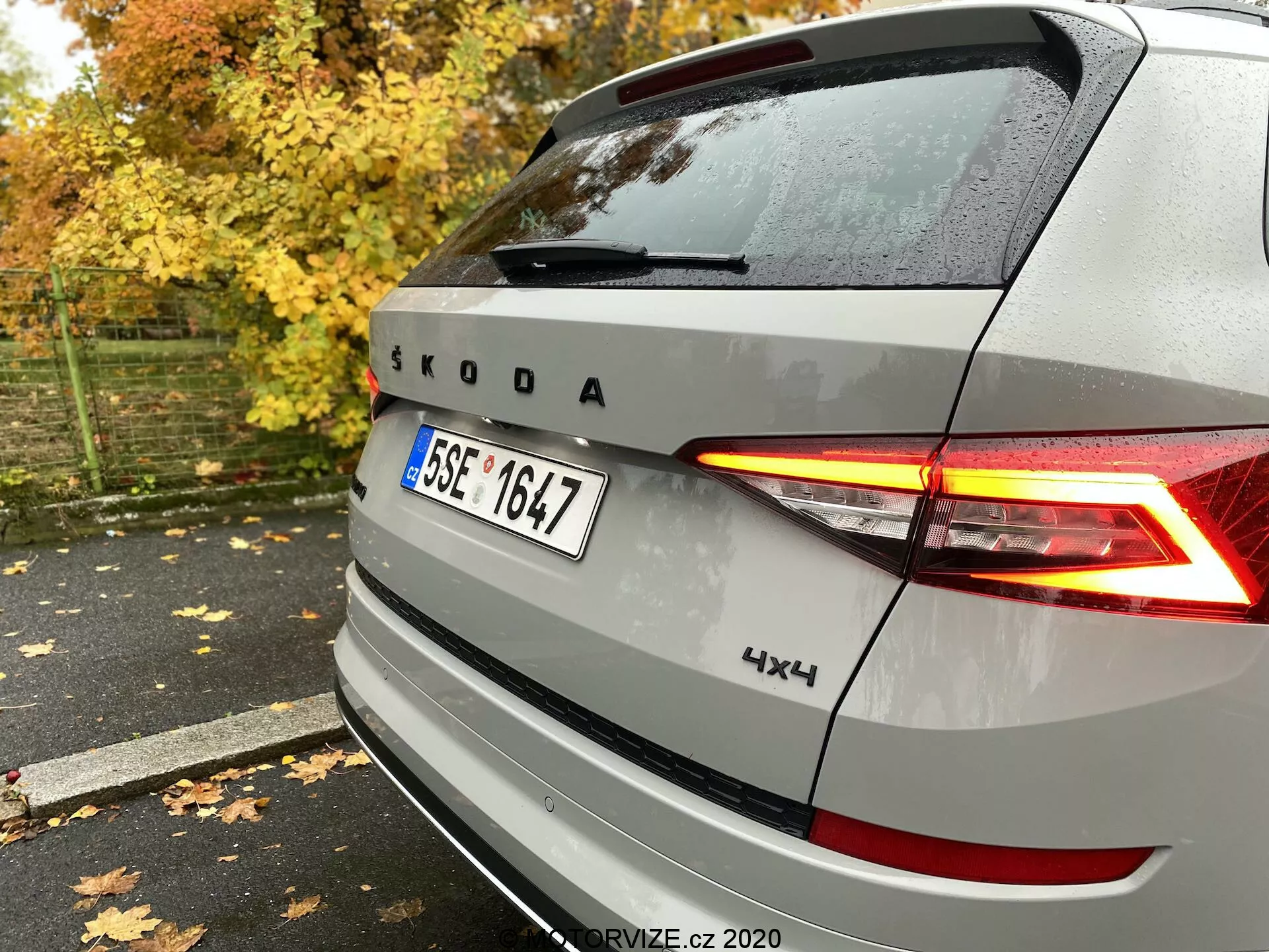 Tylny widok srebrnego pojazdu Škoda z podświetlanymi tylnymi światłami LED w kształcie litery C, emblematem i marką Škoda, z oznaczeniem 4x4, uchwycony z tyłu po prawej stronie na mokrym chodniku wśród jesiennych liści i drzew.