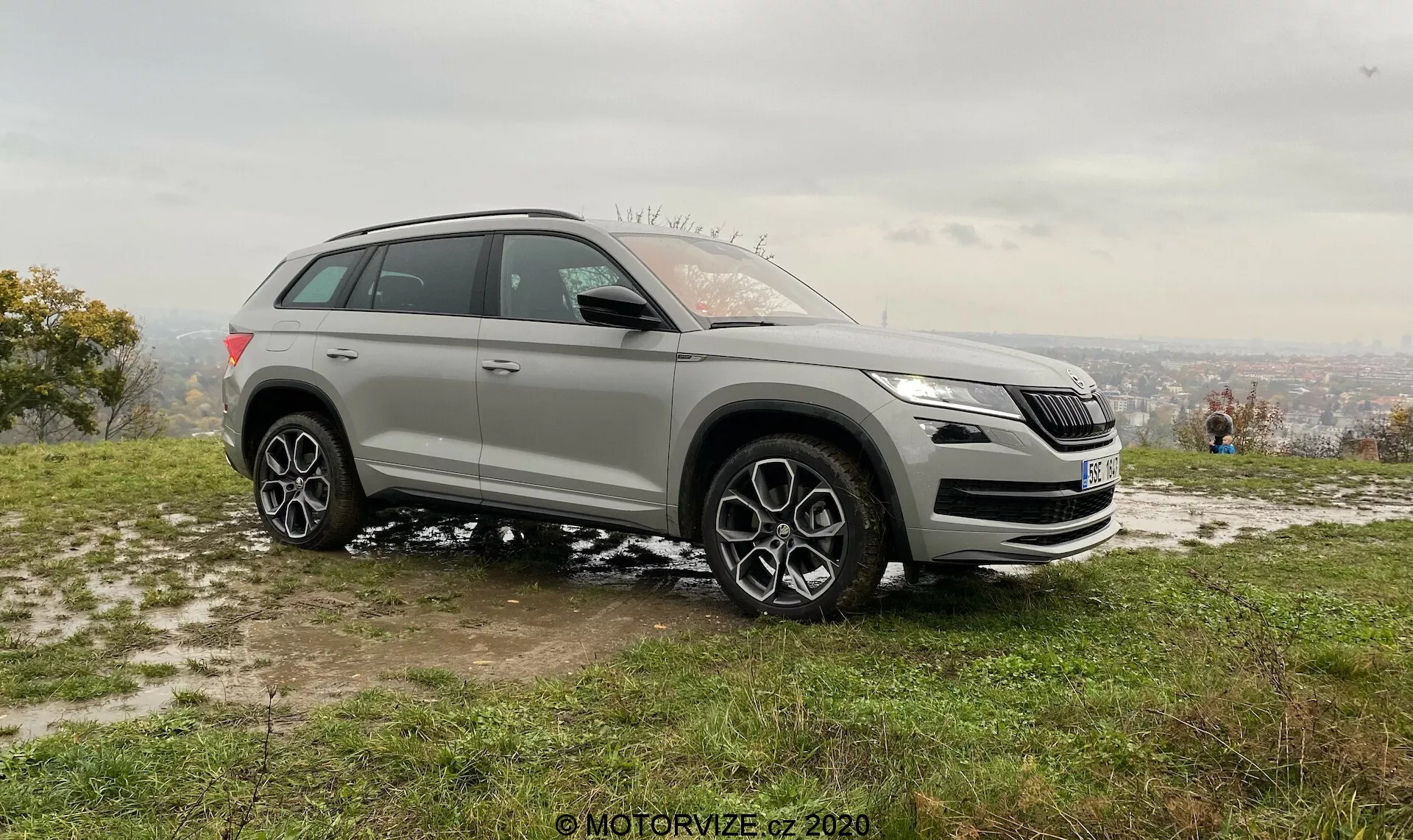 Srebrny SUV Skoda Kodiaq z 2017 roku zaparkowany na trawiastym, lekko błotnistym terenie, uchwycony z kąta bocznego przedniego. Wyraźny widok na przedni grill, reflektory, lewy profil boczny, elegancki design reflektorów z technologią LED, charakterystyczny design grilla, poniżej grilla mogą być widoczne czujniki systemów wspomagania, lusterka boczne z kierunkowskazami i możliwą detekcją martwego pola, duże, wieloszprychowe alufelgi, opony odpowiednie do zarówno drogowych, jak i umiarkowanych warunków terenowych, sugerujące napęd na cztery koła. Umieszczony na pochyłości z zamglonym widokiem na miasto w tle pod zachmurzonym niebem, miękkie oświetlenie sugerujące wczesny poranek lub późne popołudnie. Ludzie w tle cieszący się widokiem, podkreślający rekreacyjny i stylowy kontekst życia.