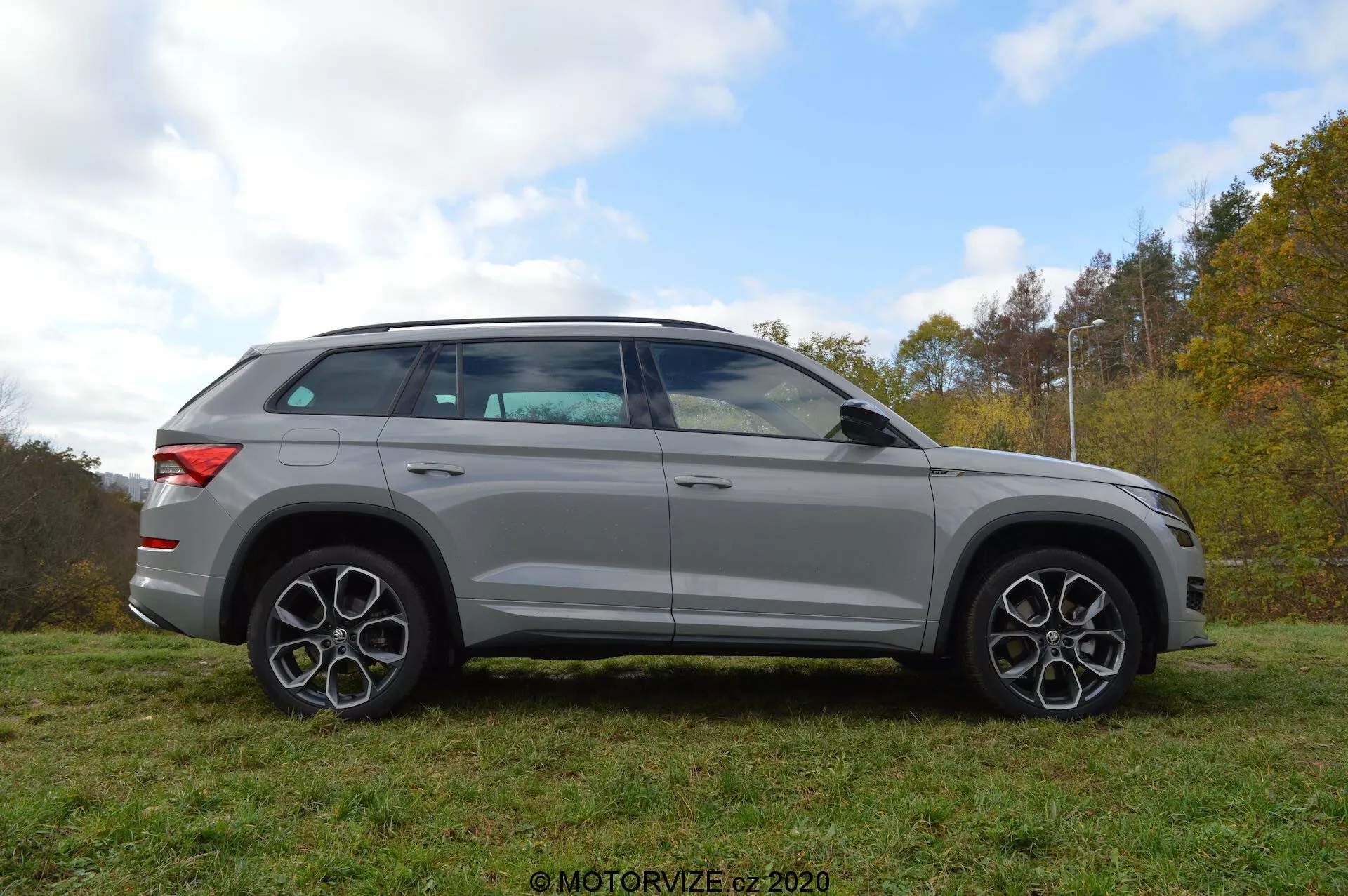Pełny profil boczny srebrnego SUV-a Skoda Kodiaq 2017 zaparkowanego na trawie z drzewami i zachmurzonym niebem w tle. Design pojazdu charakteryzuje się wyrazistymi liniami, dużymi felgami aluminiowymi i charakterystycznymi, wieloramiennymi kołami. Lusterka, które mają wbudowane kierunkowskazy, są zamontowane tuż pod linią okien. Szyby są lekko przyciemniane, a linia dachu delikatnie opada ku tyłowi, co wskazuje na sportowy profil zbliżony do coupé. SUV jest również wyposażony w system relingów dachowych.