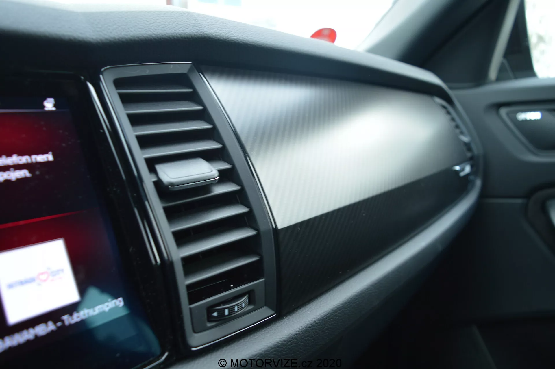 Szczegół wnętrza Skoda Kodiaq 2017: Widok z perspektywy pasażera, skupiający się na obszarze deski rozdzielczej przednim pasażerem z nawiewem powietrza, wykończeniem ze szczotkowanego metalu, teksturą w stylu włókna węglowego, częściowo widocznym ekranem systemu infotainment, wskaźnikiem poduszki powietrznej pasażera oraz oświetleniem ambientowym.