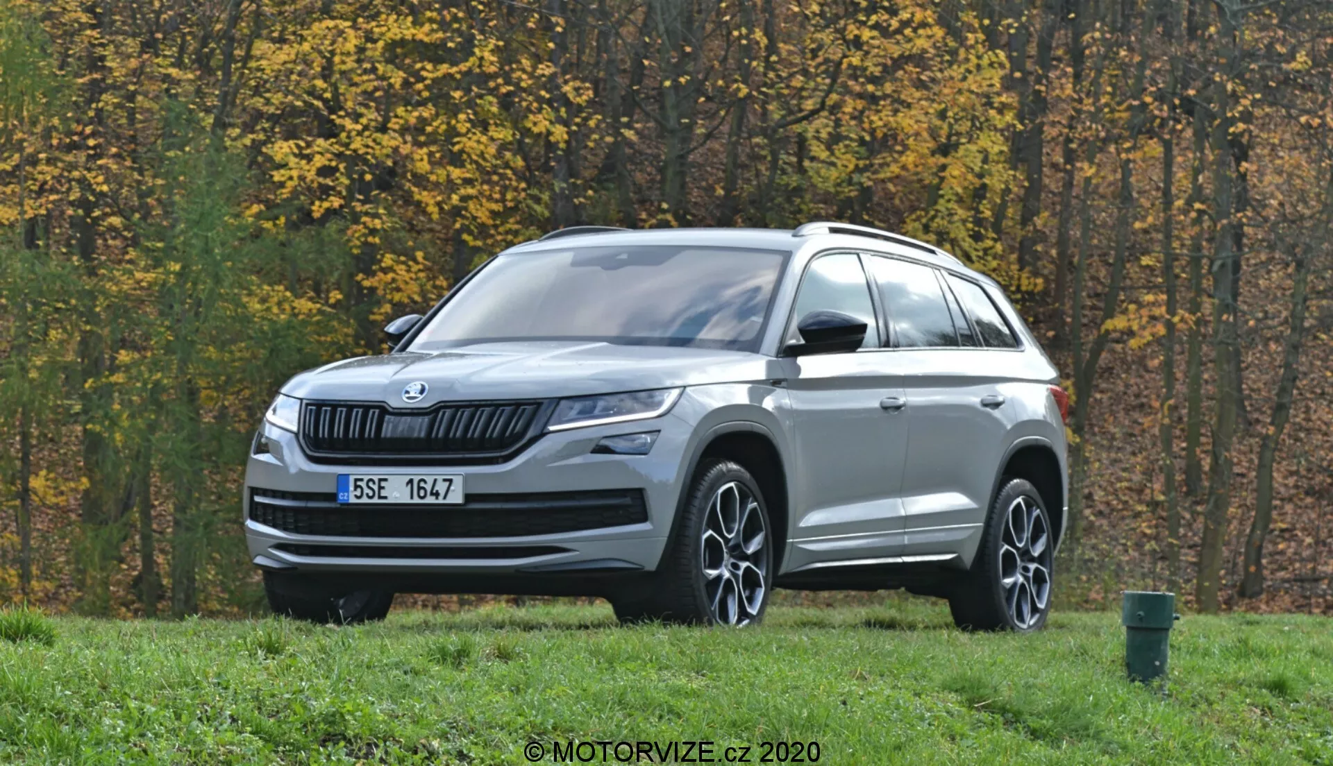 Obraz przedstawia SUV-a Skoda Kodiaq 2017 zaparkowanego na obszarze trawiastym z jesiennymi drzewami w tle. Sfocony pod kątem trzech czwartych od przodu, pojazd ma poziomą kratkę wlotu powietrza z logo Skoda w centrum. Eleganckie reflektory LED rozciągają się na boki, towarzyszą im światła przeciwmgielne na dolnej części zderzaka. Maska ma subtelne linie zagięć, a dolna część przedniego zderzaka jest ozdobiona srebrnym wykończeniem. Profil pojazdu zawiera wieloramienne felgi aluminiowe, lusterka boczne z wbudowanymi kierunkowskazami, relingi dachowe i rzeźbione linie nadwozia. Czujniki lub kamery na dolnej części zderzaka sugerują zaawansowane systemy wspomagania kierowcy (ADAS). Otoczenie na świeżym powietrzu obejmuje asfaltową drogę i zielony kosz na śmieci na prawej krawędzi kadru.