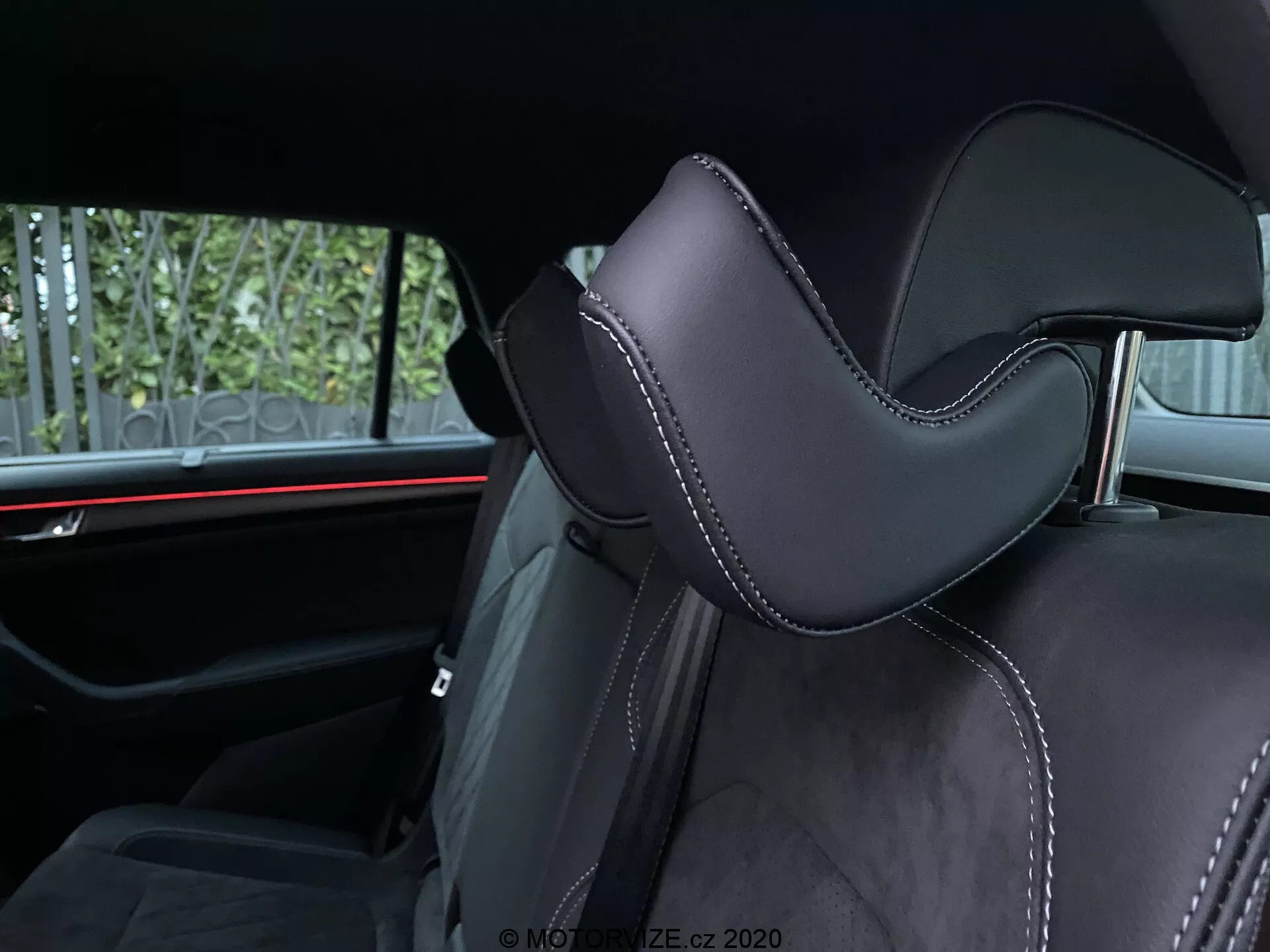 Wnętrze samochodu Skoda Kodiaq 2017 skoncentrowane na siedzeniu. Ujęcie pod kątem podkreśla część zagłówka, tapicerowaną czarnym materiałem, prawdopodobnie skórą lub wysokiej jakości syntetykiem, z wyraźnym konturem dla wygody. Precyzyjne szwy z kontrastującą nicią dodają luksusowych akcentów. W tle widoczne jest poziome czerwone oświetlenie ambientowe, które podkreśla nowoczesne elementy wzornictwa motoryzacyjnego. Widoczne udogodnienia dla pasażerów tylnych to pasy bezpieczeństwa i być może środkowy podłokietnik, co wskazuje na drugi rząd siedzeń.