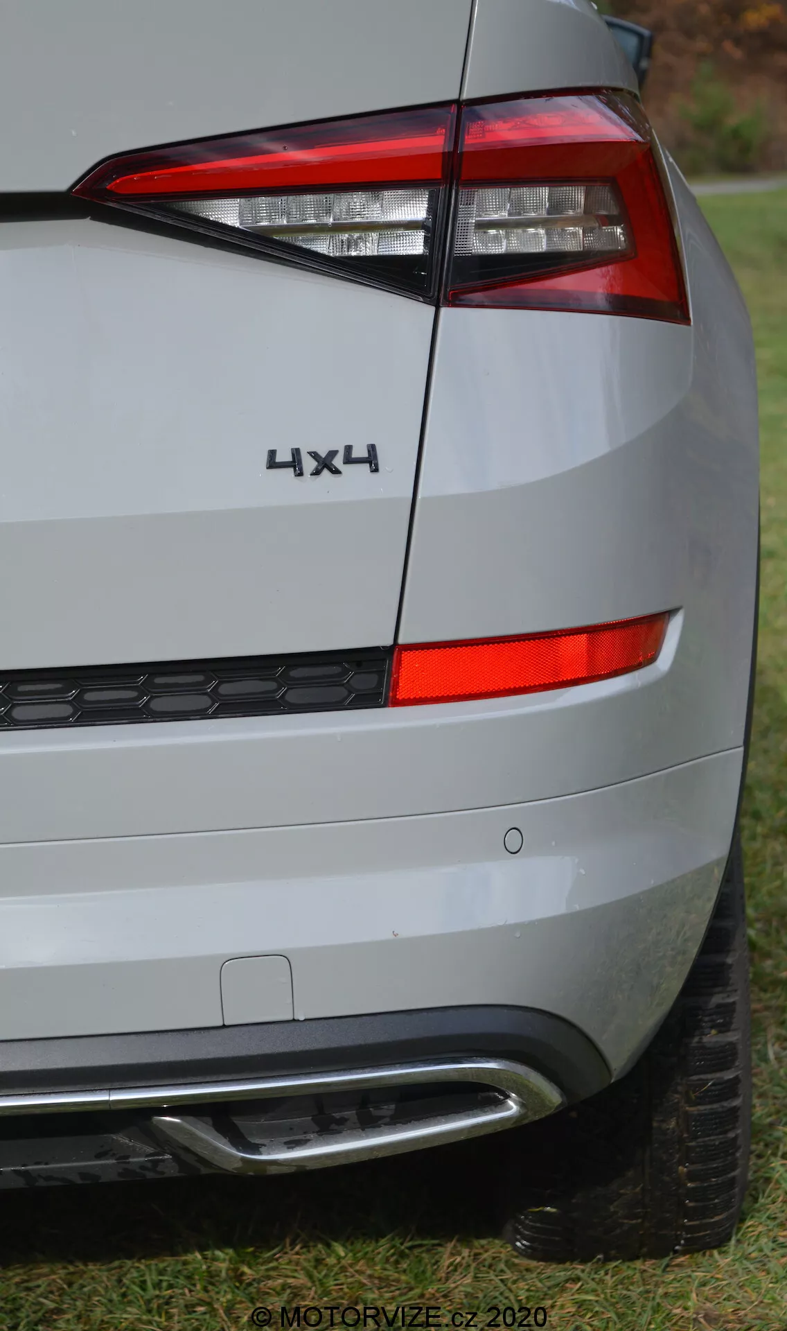 Zbliżenie na lewą tylną część Skody Kodiaq z 2017 roku. ZdjÄ™cie koncentruje siÄ™ na tylnym Å›wietle, które wyposaÅ¼ono w nowoczesne Å›wiatÅ‚a LED o ostrym, kÄ…towym wzornictwie i wyraÅºnych podziaÅ‚ach dla róÅ¼nych funkcji Å›wiateÅ‚. Symbol '4X4' wskazuje na napÄ™d na cztery koÅ‚a. Tylny zderzak zawiera odblaski i czujniki, prawdopodobnie do asysty w parkowaniu, oraz widoczny wydech. Lakier metalicznie srebrny i subtelne linie nadwozia sÄ… widoczne, a tÅ‚o stanowi trawa i drzewa, co sugeruje otoczenie podmiejskie na Å›wieÅ¼ym powietrzu. Znak wodny 'MOTORVIZE.cz 2020'.