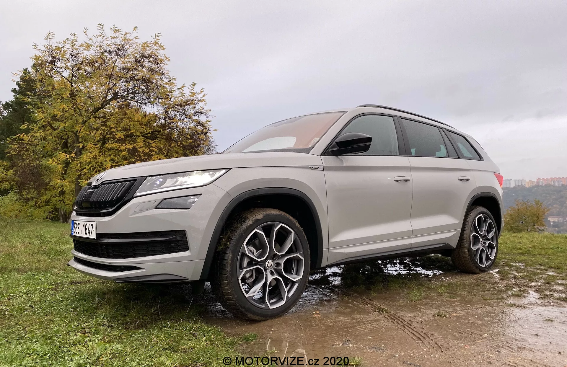 Przedni widok trzy czwarte SUV-a Škoda Kodiaq 2017 wykonany z lekko niskiego kąta, podkreślający jego wysokość i postawę. Pojazd zaparkowany jest na nierównej powierzchni z trawy i błota, co uwydatnia jego zdolności terenowe. Škoda Kodiaq charakteryzuje się wyraźnym grillem i ostrymi reflektorami LED. Precyzyjny design obejmuje duże, wieloramienne felgi aluminiowe, wysoki prześwit i opony na każdą pogodę. Nadwozie jest pomalowane na neutralny metaliczny kolor z przyciemnianymi szybami, a chromowane akcenty zwiększają jego luksusowy wygląd. Nowoczesne funkcje obejmują lusterka boczne w kolorze nadwozia z wbudowanymi kierunkowskazami i prawdopodobnie systemami ostrzegania o martwym polu.