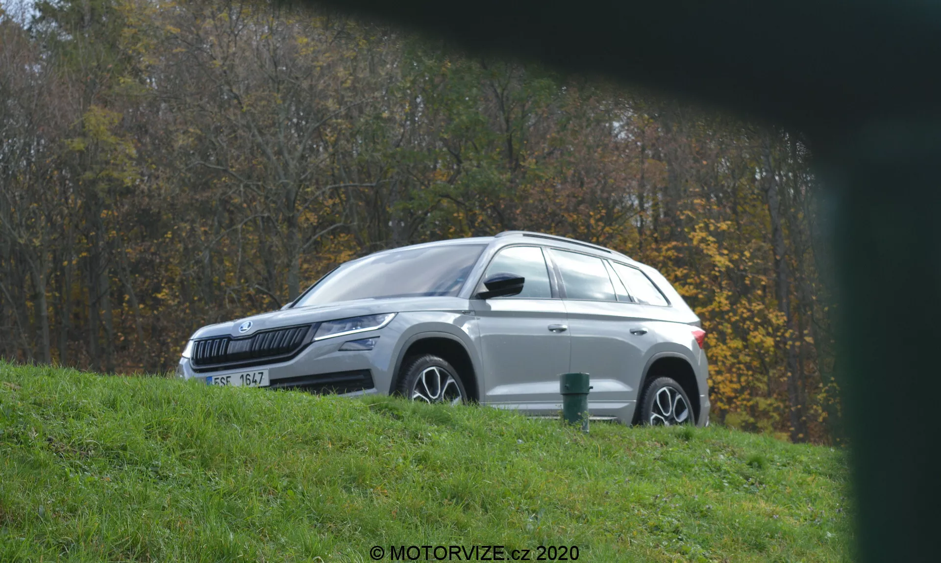 Widok z boku na białe SUV Skoda Kodiaq, prawdopodobnie nowoczesny model z lat 2020, sfotografowany z lekko podwyższonego kąta, zaparkowany na trawie obok utwardzonej drogi w czasie jesieni, z pochmurnym niebem; ukazujący zamknięte drzwi, srebrne relingi dachowe i jesienne liście w tle oraz zawierający znak wodny 'MOTORVIZE.cz 2020'.