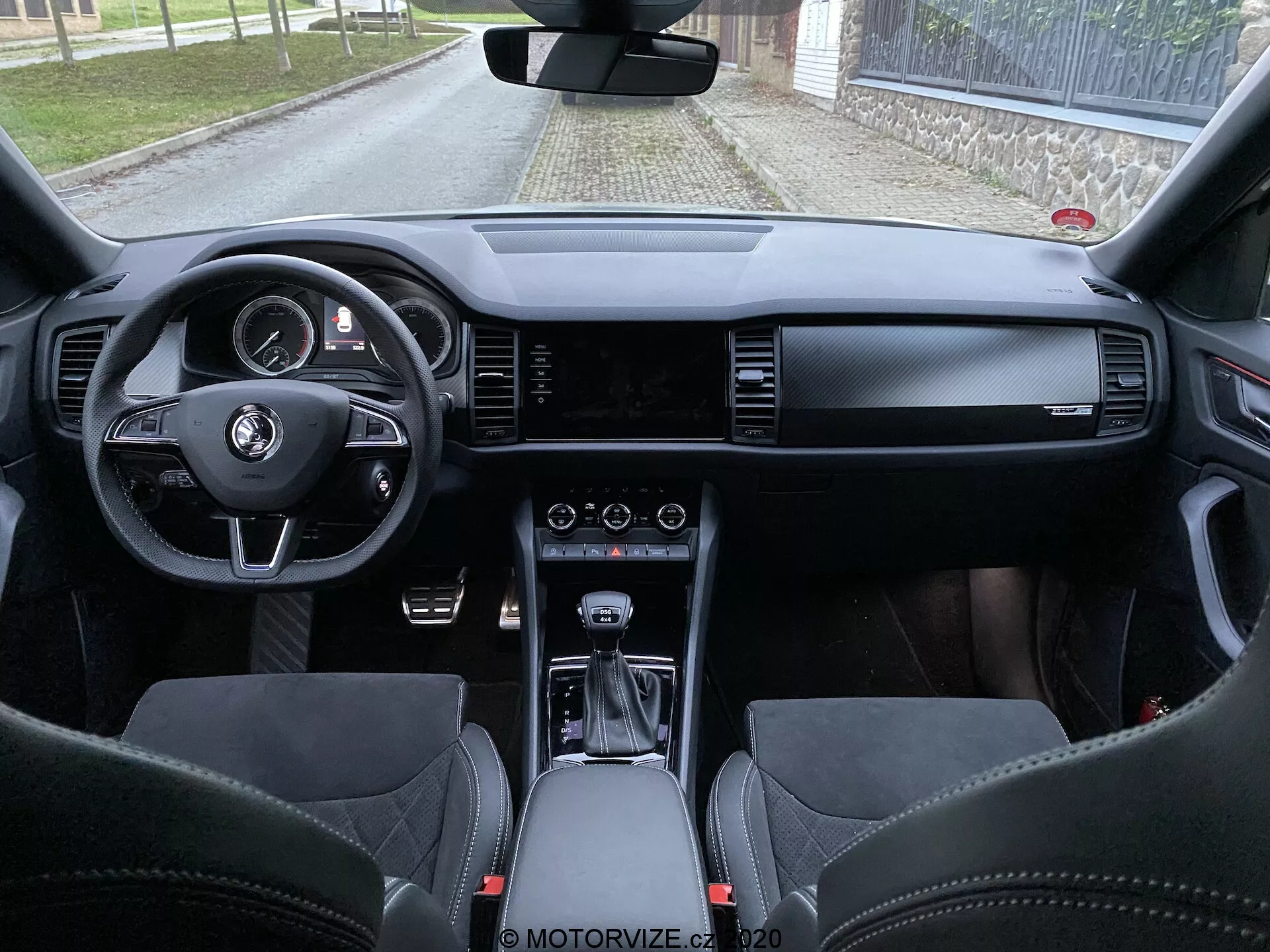 Widok wnętrza Skoda Kodiaq 2017 z przestrzeni tylnego siedzenia, ukazującego deskę rozdzielczą, kierownicę z przyciskami wielofunkcyjnymi, cyfrowy zestaw wskaźników, szeroki ekran informacyjno-rozrywkowy, kontrolki HVAC, selektor automatycznej skrzyni biegów, wysokiej jakości skórzane materiały i sportowe siedzenia z zintegrowanymi zagłówkami.