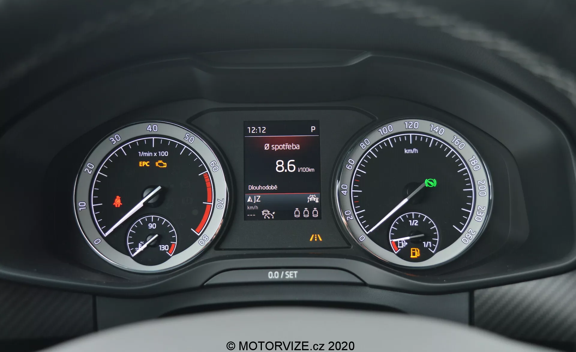 Widok z perspektywy kierowcy na deskę rozdzielczą Škoda Kodiaq 2017, z nowoczesnym i czystym designem łączącym wyświetlacze analogowe i cyfrowe. Po lewej: analogowy obrotomierz i wskaźnik poziomu paliwa, w środku: cyfrowy wyświetlacz wielofunkcyjny pokazujący godzinę (12:12), bieg (P), prędkość (8,6 km/h), licznik kilometrów (15,666 km) i nieokreślone ikony. Po prawej: analogowy prędkościomierz z wskaźnikiem poziomu naładowania baterii. W sumie deska rozdzielcza jest głównie ciemna z białym podświetleniem.