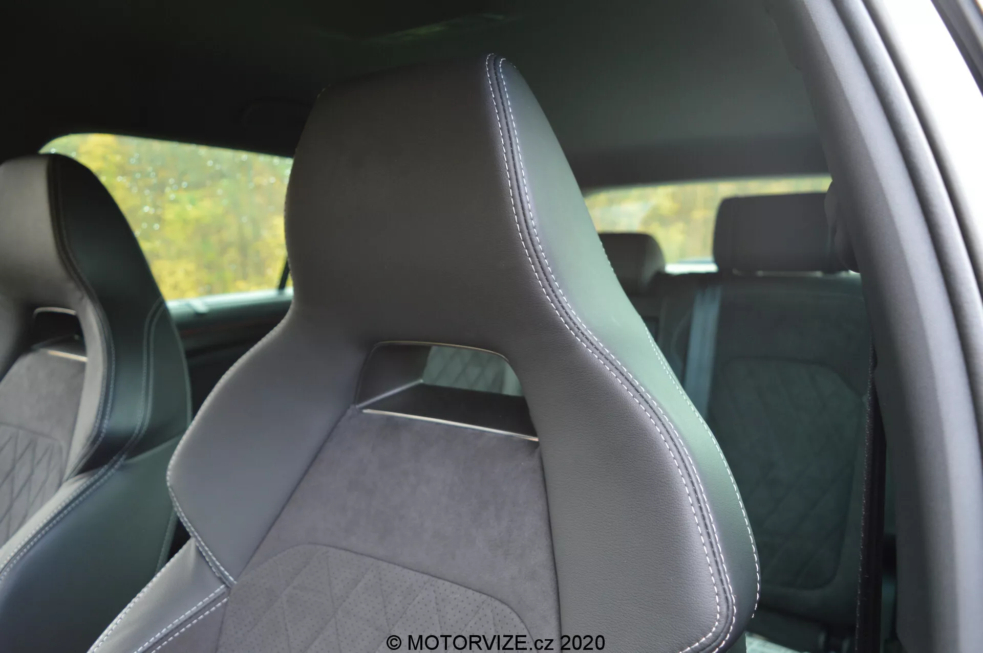 Wnętrze Škoda Kodiaq 2017, skupiające się na przednim fotelu kierowcy z tylnego bocznego perspektywy, podkreślające tapicerkę ze skóry i teksturowanej tkaniny z precyzyjnymi przeszyciami na krawędziach. Nowoczesne, sportowe siedzisko posiada wyraźnie zaznaczone boczne wzmocnienia dla lepszego podparcia, zintegrowany zagłówek i wzór przeszyć w kształcie diamentu, co sugeruje prestiżową estetykę. Tło wnętrza jest nieco rozmyte, aby podkreślić siedzisko.