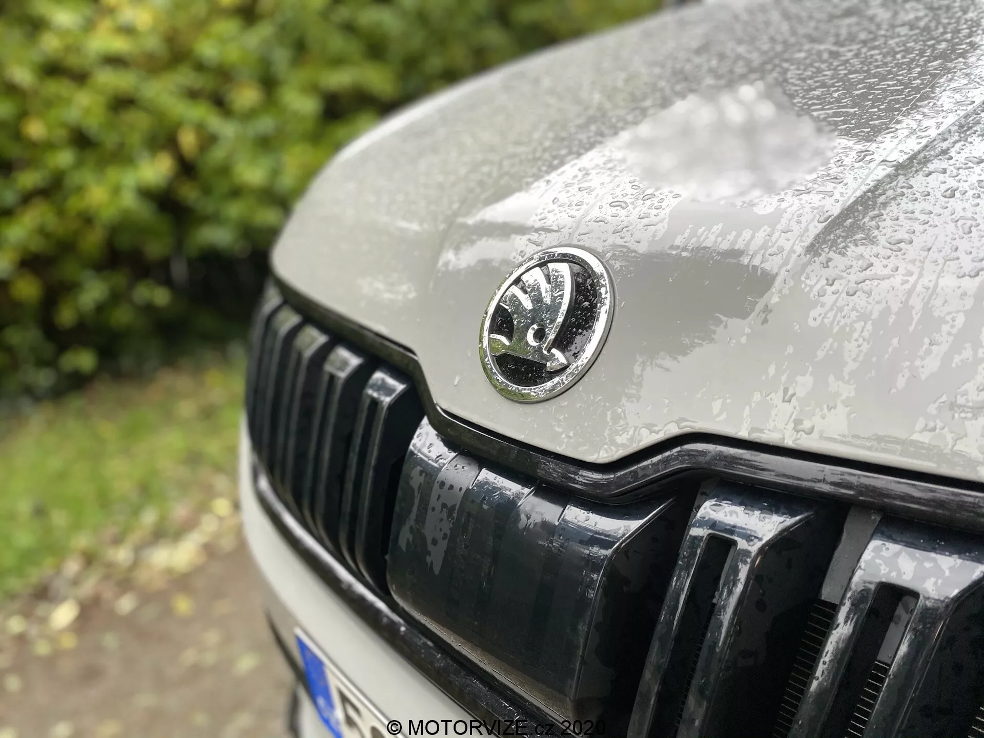 Zbliżenie przedniej części samochodu Skoda Kodiaq 2017, koncentrujące się głównie na grillu i emblemacie producenta. Okrągły emblemat znajduje się w centrum, ukazując delikatne szczegóły z kroplami wody wskazującymi na niedawny deszcz. Lekko odchylony od środka kąt ukazuje maskę i błyszczące czarne pionowe żaluzje grillu w ostrym zbliżeniu z małą głębią ostrości.