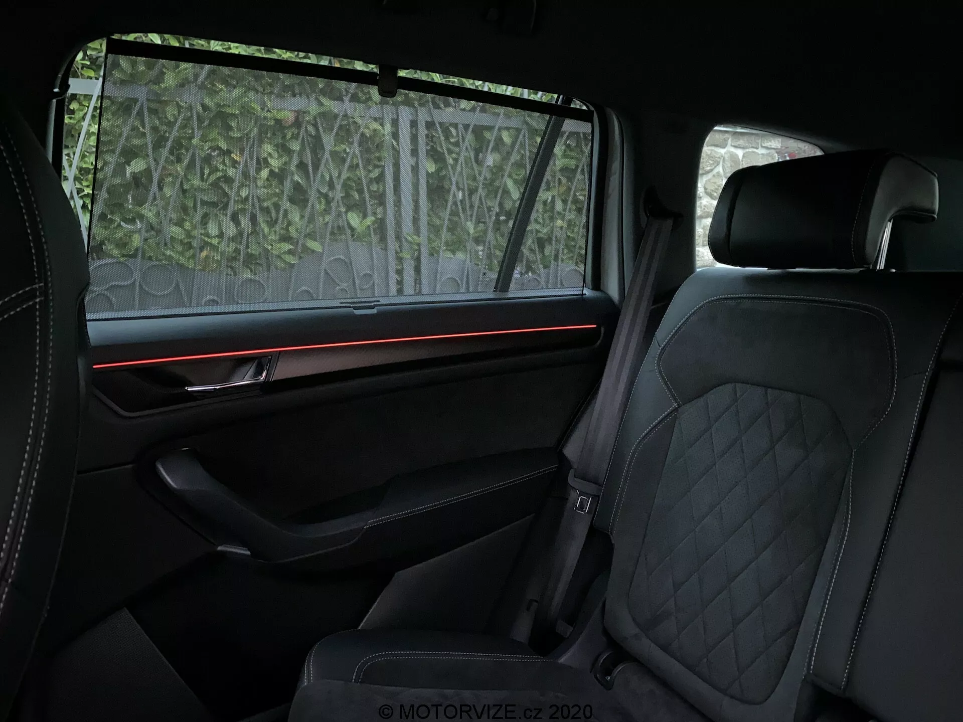 Widok wnętrza Skody Kodiaq 2017 skupiający się na tylnym przedziale pasażerskim, ukazujący luksusowe elementy, takie jak pikowane skórzane tapicerki na przednich i tylnych siedzeniach, czerwona ambientowa listwa świetlna na dobrze wyposażonym wyściełanym panelu drzwi z premium wykończeniem oraz częściowo przezroczystą roletką przeciwsłoneczną w oknie. Nacisk kładziony jest na komfort i elementy projektowania najwyższej jakości.