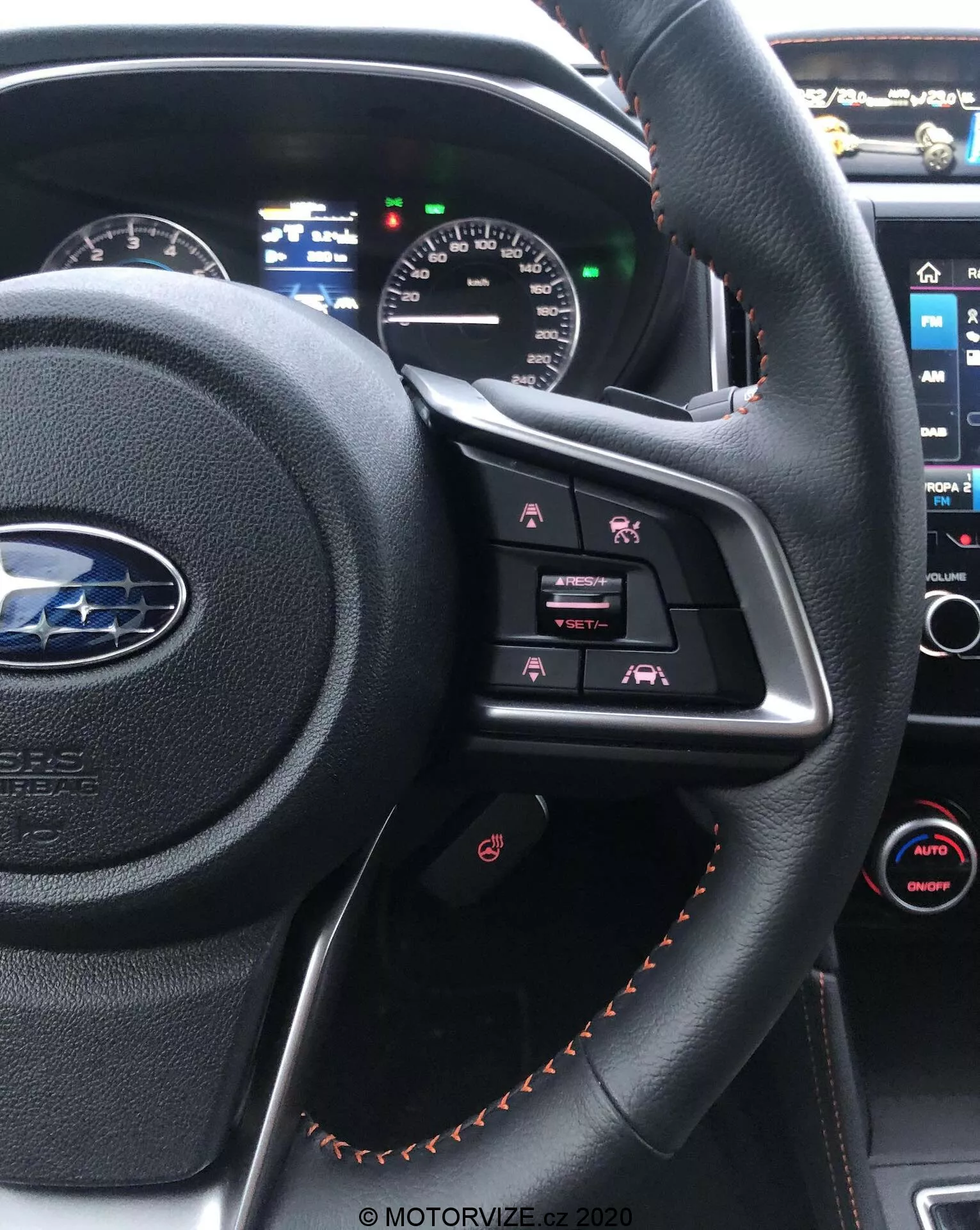 Widok wnętrza Subaru XV 2018 z fotela kierowcy, podkreślający kierownicę obszytą czarną skórą z pomarańczowymi szwami, z przyciskami sterowania multimediami i częściowo widocznym panelem instrumentów z analogowymi i cyfrowymi wyświetlaczami. W tle widoczna jest także konsola centralna z ekranem dotykowym systemu multimedialnego i dwustrefowym automatycznym sterowaniem klimatyzacją.