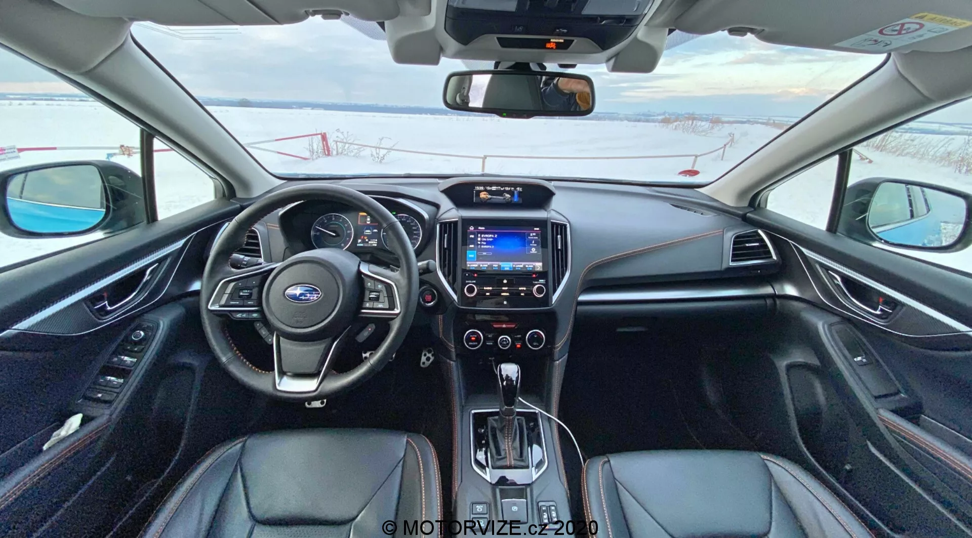 Widok wnętrza Subaru XV 2018 (Subaru XV 1.6i Trend Lineartronic) z siedzenia kierowcy przedstawiający skórzaną kierownicę z wbudowanymi kontrolami, częściowo widoczny zestaw analogowych i cyfrowych wskaźników, centralny ekran infotainmentowy z niebieskim interfejsem, pokrętła sterowania klimatem, automatyczną dźwignię zmiany biegów otoczoną przyciskami operacyjnymi, czarną tapicerkę z srebrnymi akcentami, sterowanie podgrzewaniem siedzeń i lusterko wsteczne z wyświetlaczem cyfrowym. Samochód jest zaparkowany, a na zewnątrz leży śnieg, co wskazuje na zimny klimat.