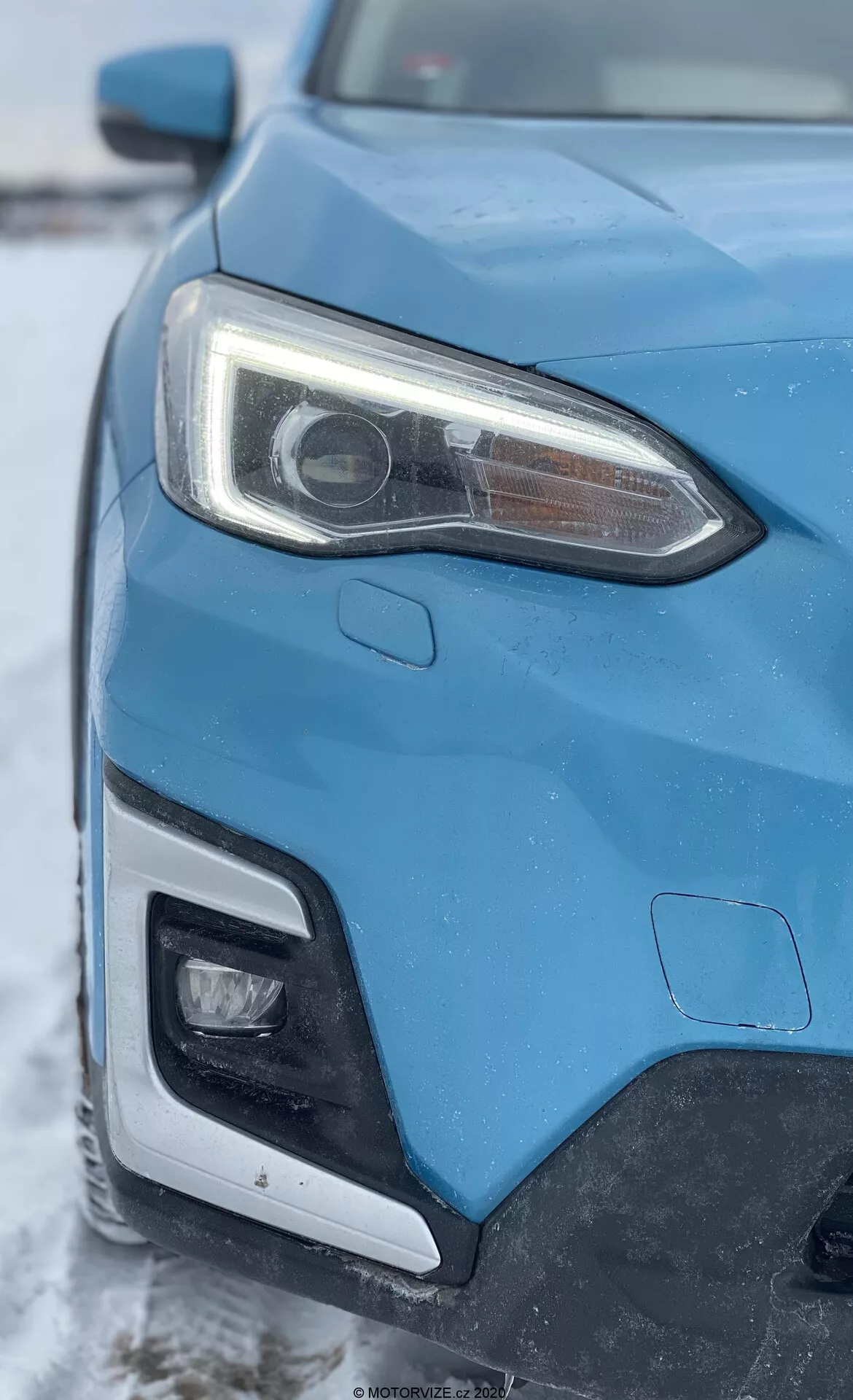 Zbliżenie lewej przedniej części niebieskiego Subaru XV 1.6i Trend Lineartronic z 2018 roku, uchwycone pod kątem ukośnym, skupiające się na reflektorze oraz części grilla i przedniego zderzaka. Wyraźny lewy reflektor posiada soczewkę projektora, co wskazuje na systemy HID lub LED oraz zintegrowane LED-owe światła do jazdy dziennej. Zdjęcie pokazuje również część przedniego zderzaka, która prawdopodobnie mieści dodatkowe oświetlenie. Karoseria samochodu wykazuje ostre linie i aerodynamiczne kształty, na tle zimowego krajobrazu z lekką warstwą śniegu.