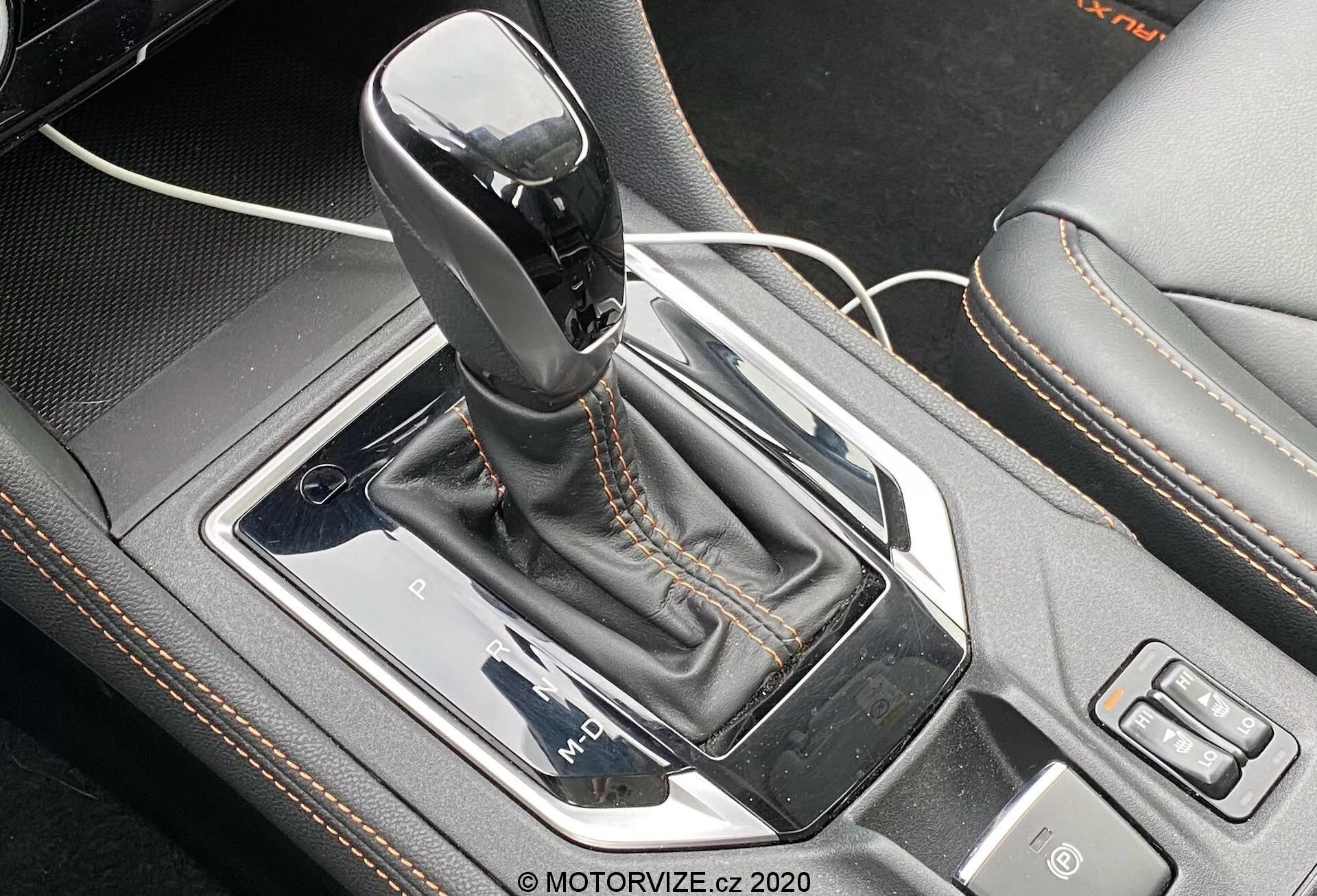 Zbliżenie na automatyczną dźwignię zmiany biegów Lineartronic w Subaru XV 2018 1.6i Trend. Dźwignia zmiany biegów, w pozycji parkowania, z lśniącą czarną górą i srebrnym akcentem, jest centralnie umieszczona na konsoli środkowej samochodu. Jest otoczona srebrnym panelem, osadzone w wysokopołyskowej, czarnej metalicznej ramce z mankietem z imitacji skóry z kontrastowymi szwami. Po lewej stronie przewód USB wskazuje na pobliski port, podczas gdy po prawej widoczne są przyciski wyboru trybu jazdy i innej funkcji. Perspektywa z góry i lekko z przodu podkreśla zawiłe szczegóły projektowe i funkcje technologiczne obszaru dźwigni zmiany biegów.