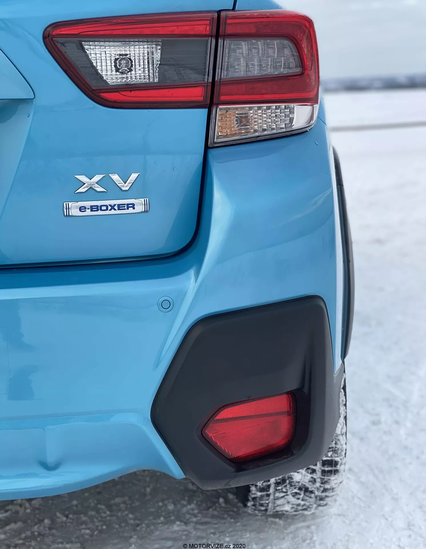Zbliżenie, tylne ujęcie niebieskiego modelu Subaru XV 2018, skupiające się na lewym tylnym narożniku od tylnego światła do tylnego koła. Główne cechy obejmują złożony zestaw tylnych świateł, oznaczenia 'XV' oraz 'e-BOXER' wskazujące na technologię hybrydową, błyszczący metaliczny niebieski lakier z czarnym plastikowym nadkolem, czarną plastikową wstawkę na tylnym zderzaku odbijającą ośnieżone podłoże sugerujące zimowy klimat oraz częściowo widoczne tylne oponę z bieżnikiem pokrytym śniegiem. Ostrość jest skupiona na tylnym narożniku, podczas gdy tło jest lekko rozmyte z powodu głębi ostrości.