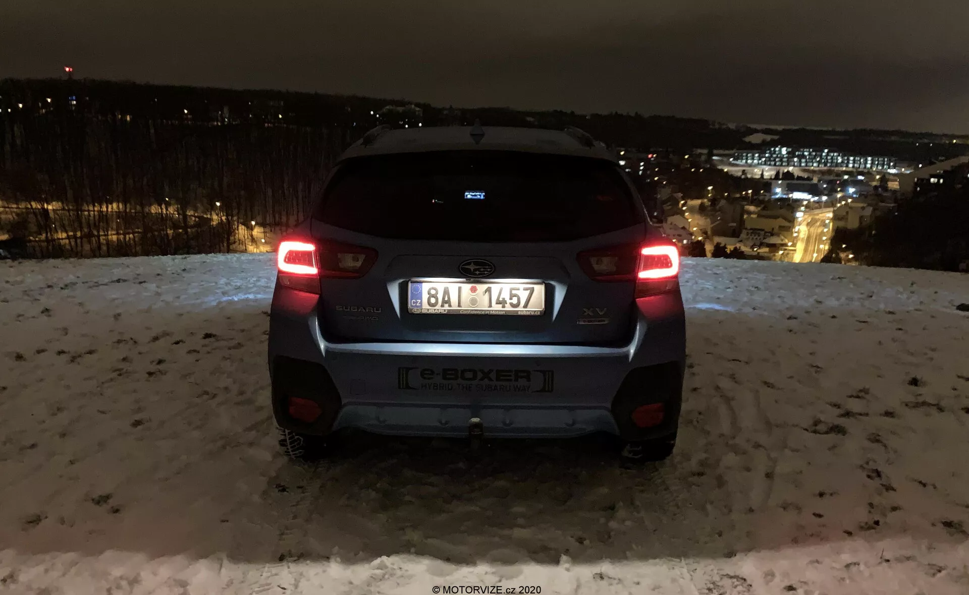 Widok tyłu Subaru XV 2018 (subaru-xv-1-6i-trend-lineartronic) uchwycony w nocy z widokiem na światła miasta w tle. Samochód znajduje się na podwyższonym terenie, pokazując oświetlone tylne światła, tylną szybę, znaczek producenta, oznaczenie modelu e-BOXER, tablicę rejestracyjną, tylny zderzak oraz kamerę lub czujnik parkowania. Odbijające światło powierzchnie i śnieg na ziemi sugerują zimowe warunki. Nowoczesny design pojazdu obejmuje mały spojler, minimalistyczną tylną wycieraczkę i dwukolorowy zderzak, podkreślając elementy oświetleniowe i projektowe marki motoryzacyjnej.