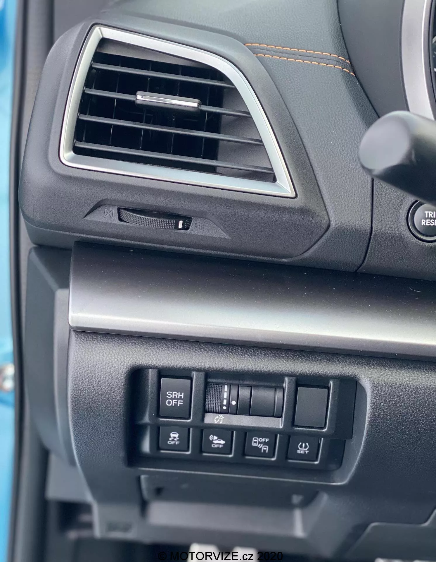 Zbliżenie na deskę rozdzielczą po stronie kierowcy w Subaru XV 1.6i Trend Lineartronic z 2018 roku. Obraz koncentruje się na prostokątnym wlocie powietrza z zaokrąglonymi krawędziami i regulatorem kierunku wlotu, częściowo widocznej kierownicy z przyciskami sterującymi dźwiękiem oraz zamkniętym schowku pod wlotem powietrza z miękką teksturą powierzchni. Widoczne są również przyciski sterujące funkcjami, takimi jak adaptacyjne reflektory (przycisk SRH OFF) i inne ustawienia pojazdu. Materiały deski rozdzielczej są wykonane z czarnego lub ciemnoszarego plastiku o różnych teksturach, co sugeruje ich trwałość i jakość.